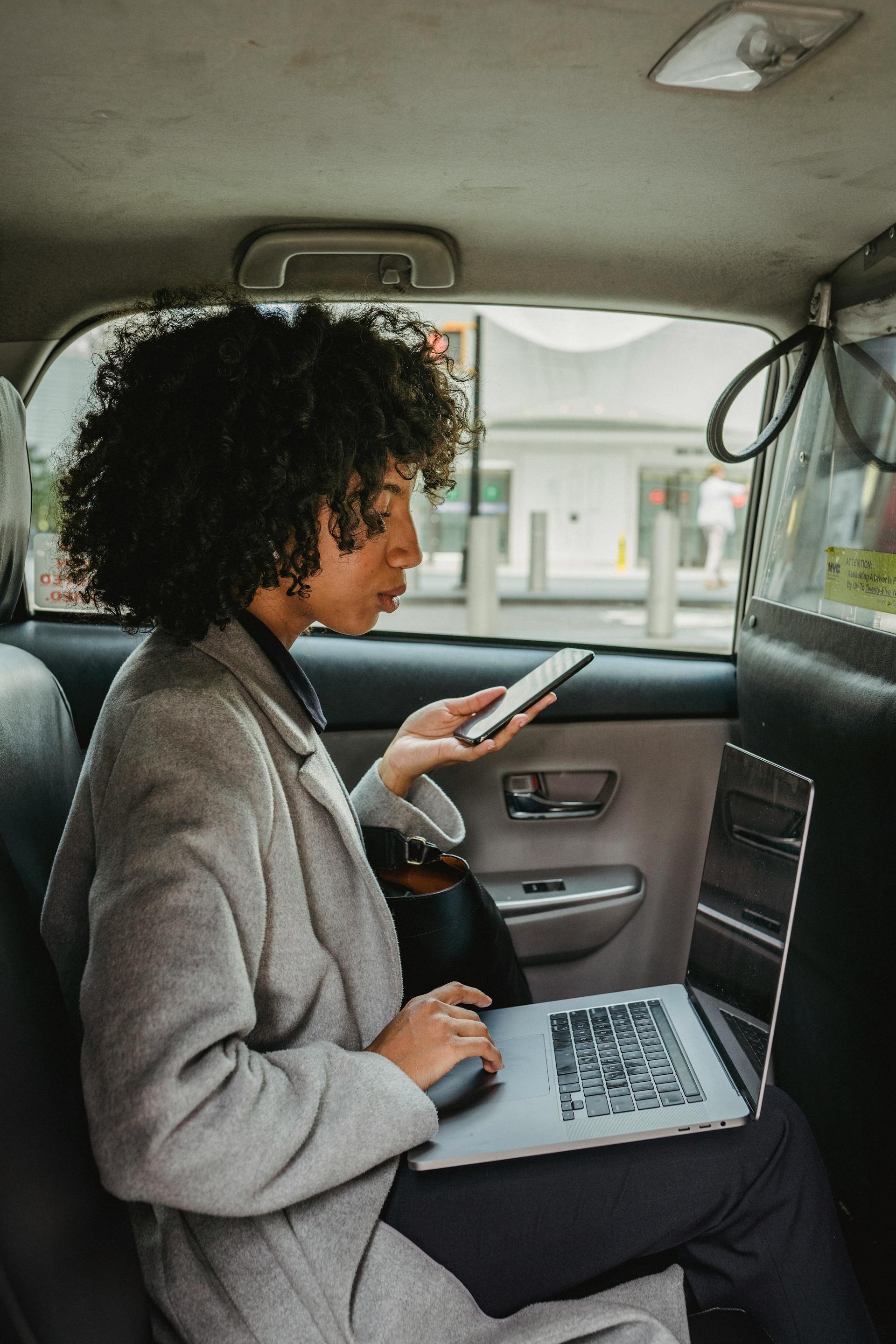 Una mujer seria sujetando un teléfono y utilizando un ordenador portátil | Fuente: Pexels