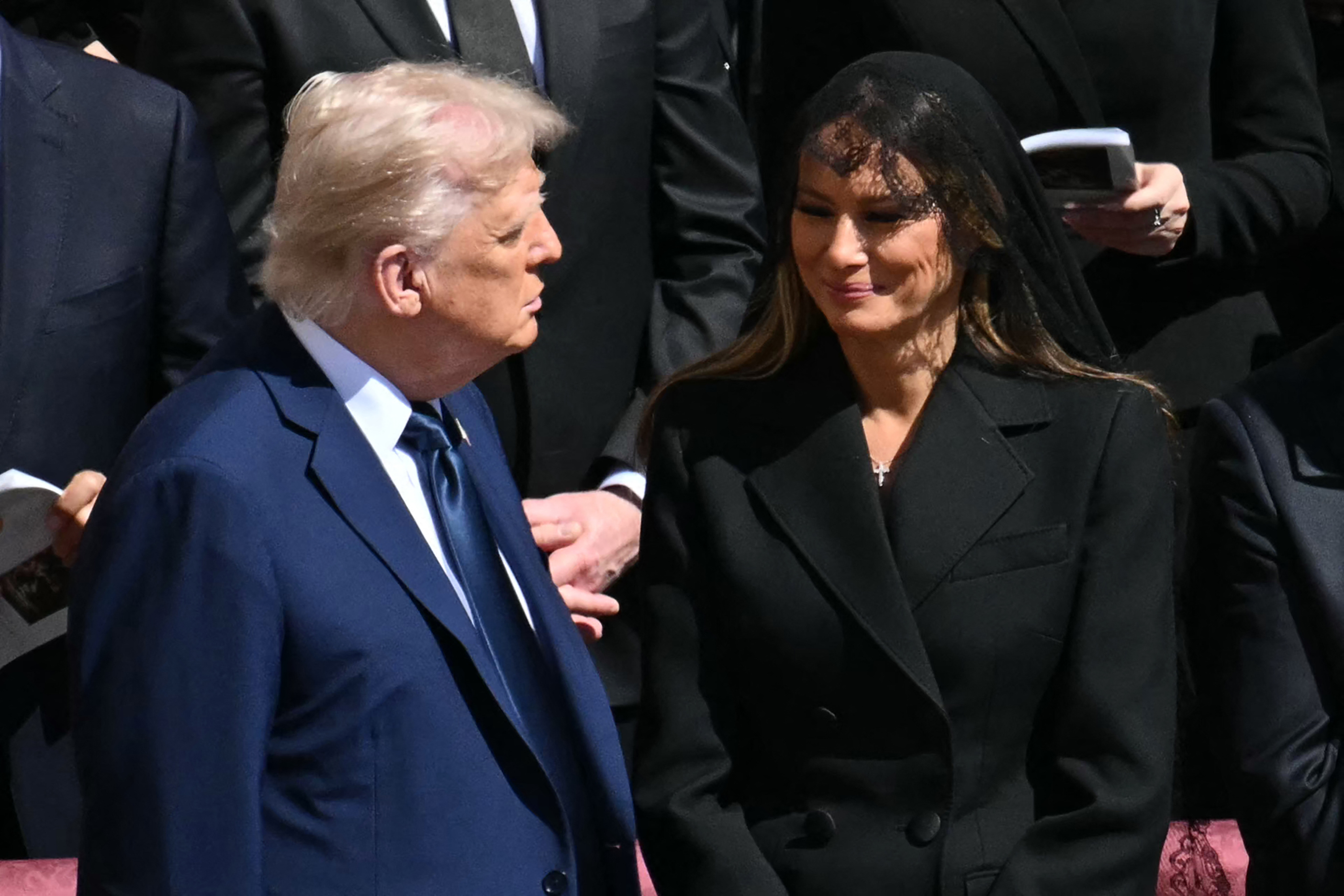 Donald y Melania están juntos, hablando. Melania luce una sutil sonrisa, de expresión suave, mientras comparten un momento de tranquila alegría en medio del luto.