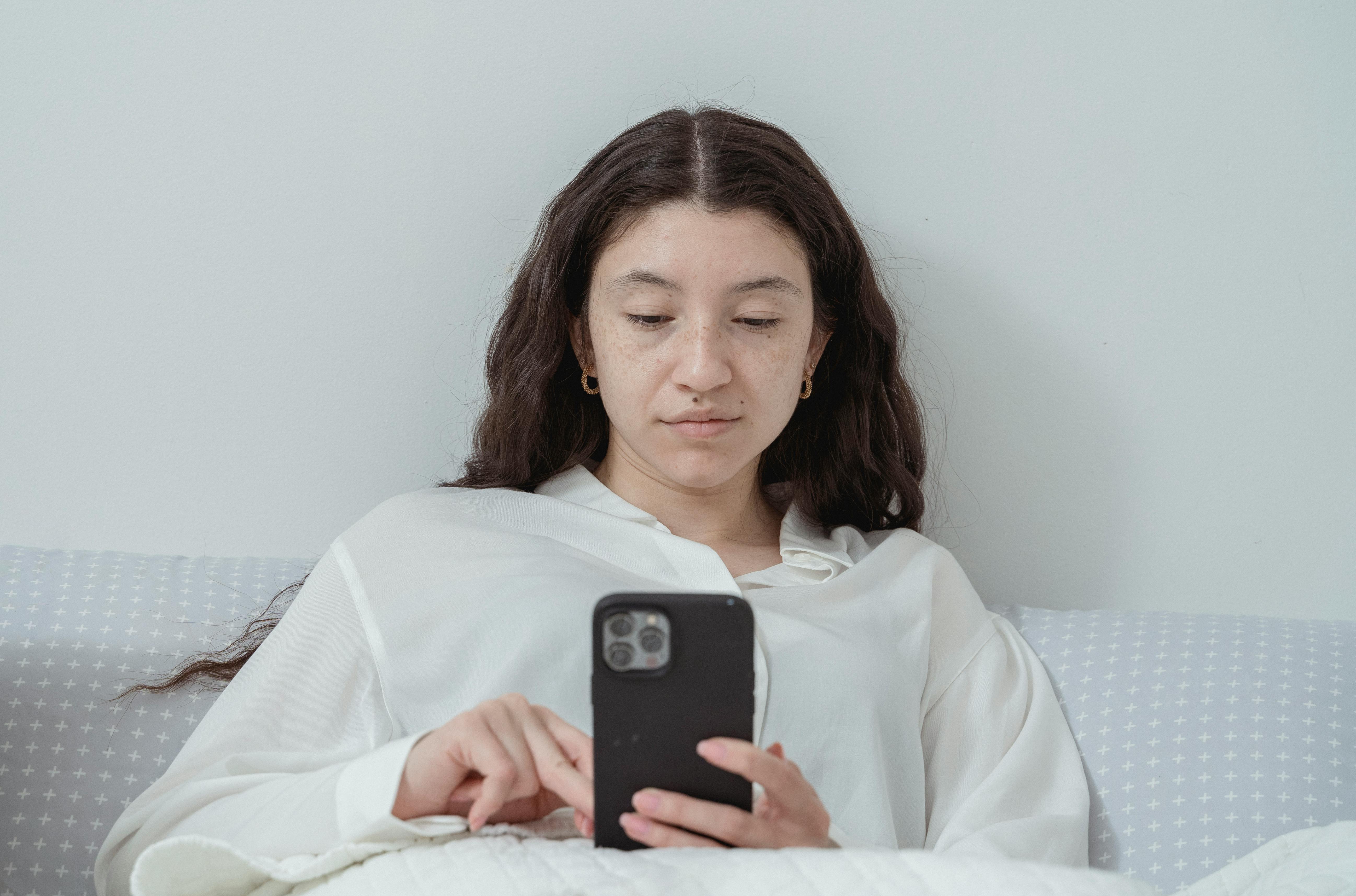 Una mujer seria utilizando su teléfono mientras está tumbada en la cama | Fuente: Pexels