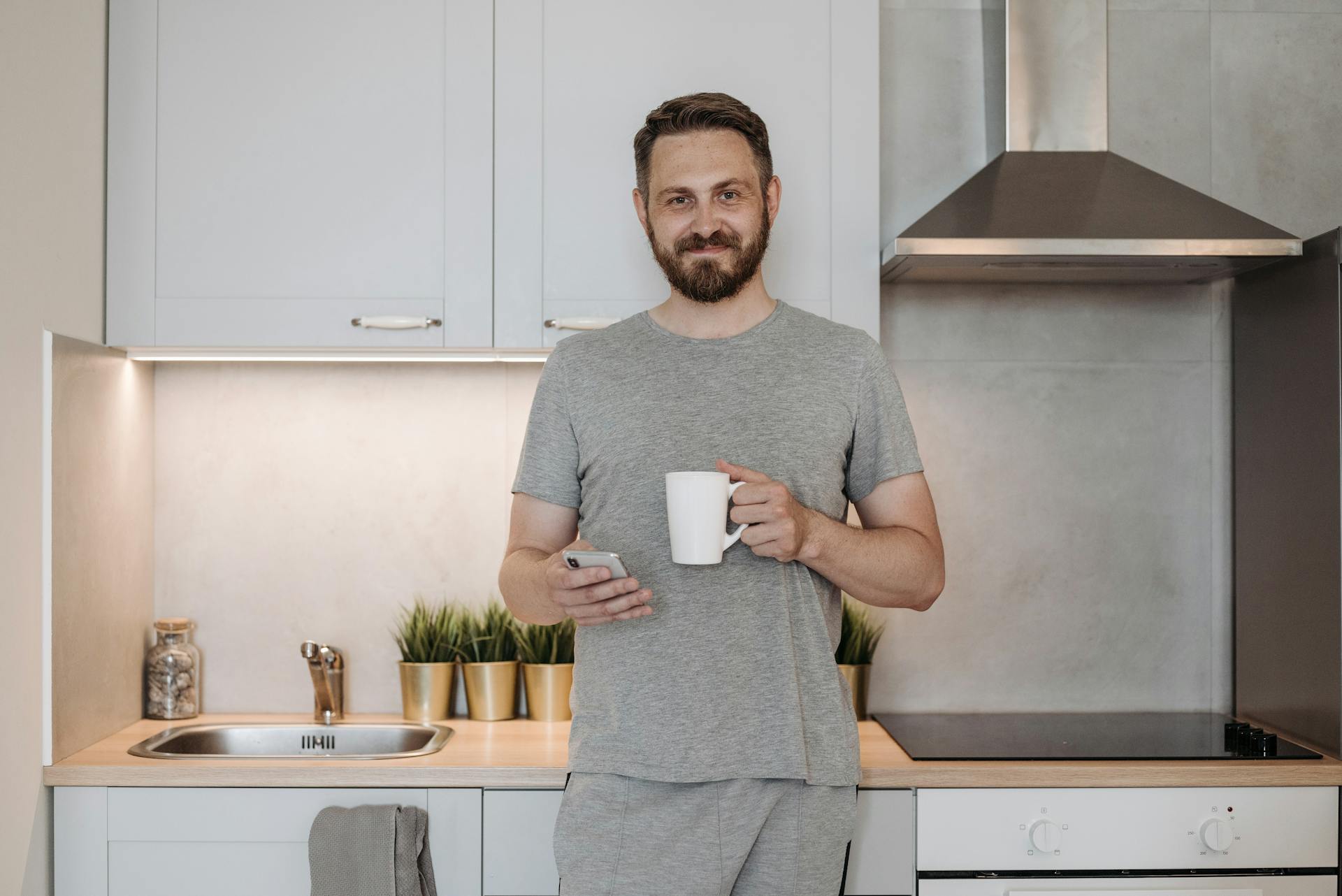 Un hombre sostiene un teléfono y una taza mientras está de pie en la cocina | Fuente: Pexels