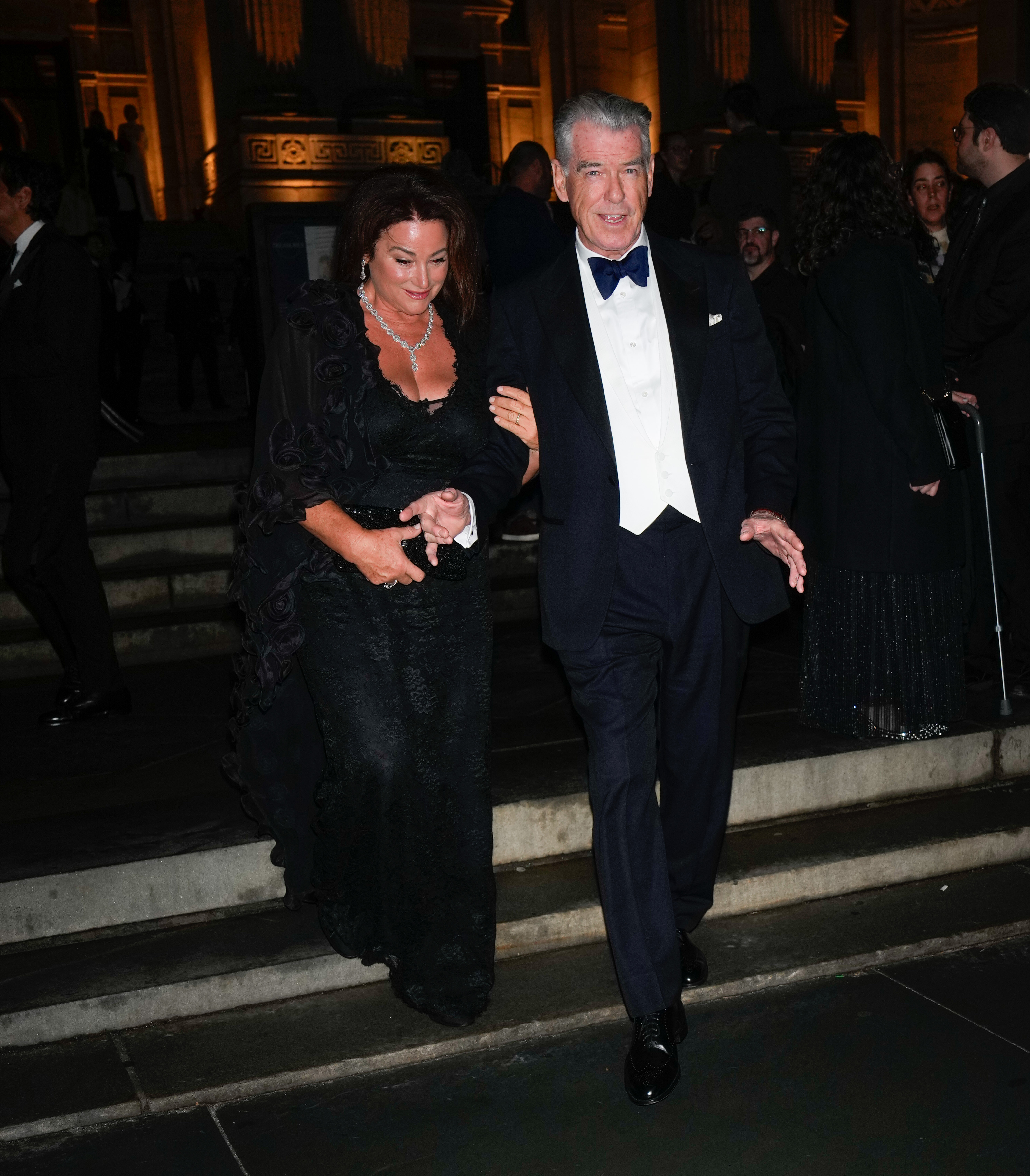 Pierce Brosnan le toma con delicadeza la mano a Keely Shaye Smith mientras bajan juntos las escaleras; Keely lleva un elegante vestido negro de encaje y joyas brillantes. | Fuente: Getty Images.