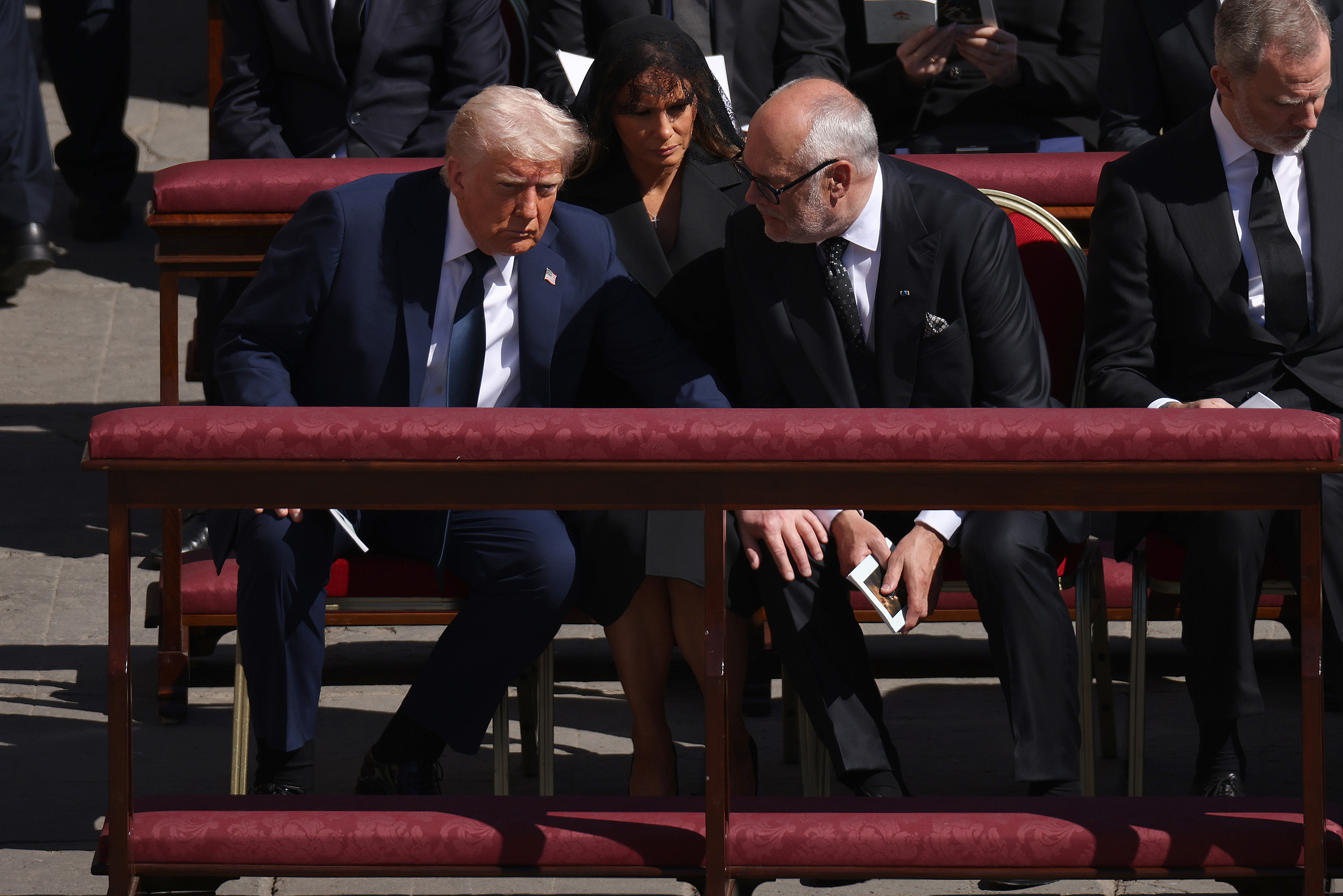 Donald y el presidente estonio Alar Karis se inclinan hacia delante en una conversación tranquila durante el funeral. Mientras tanto, Melania se sienta tranquilamente, con una postura serena, mientras observa la interacción.