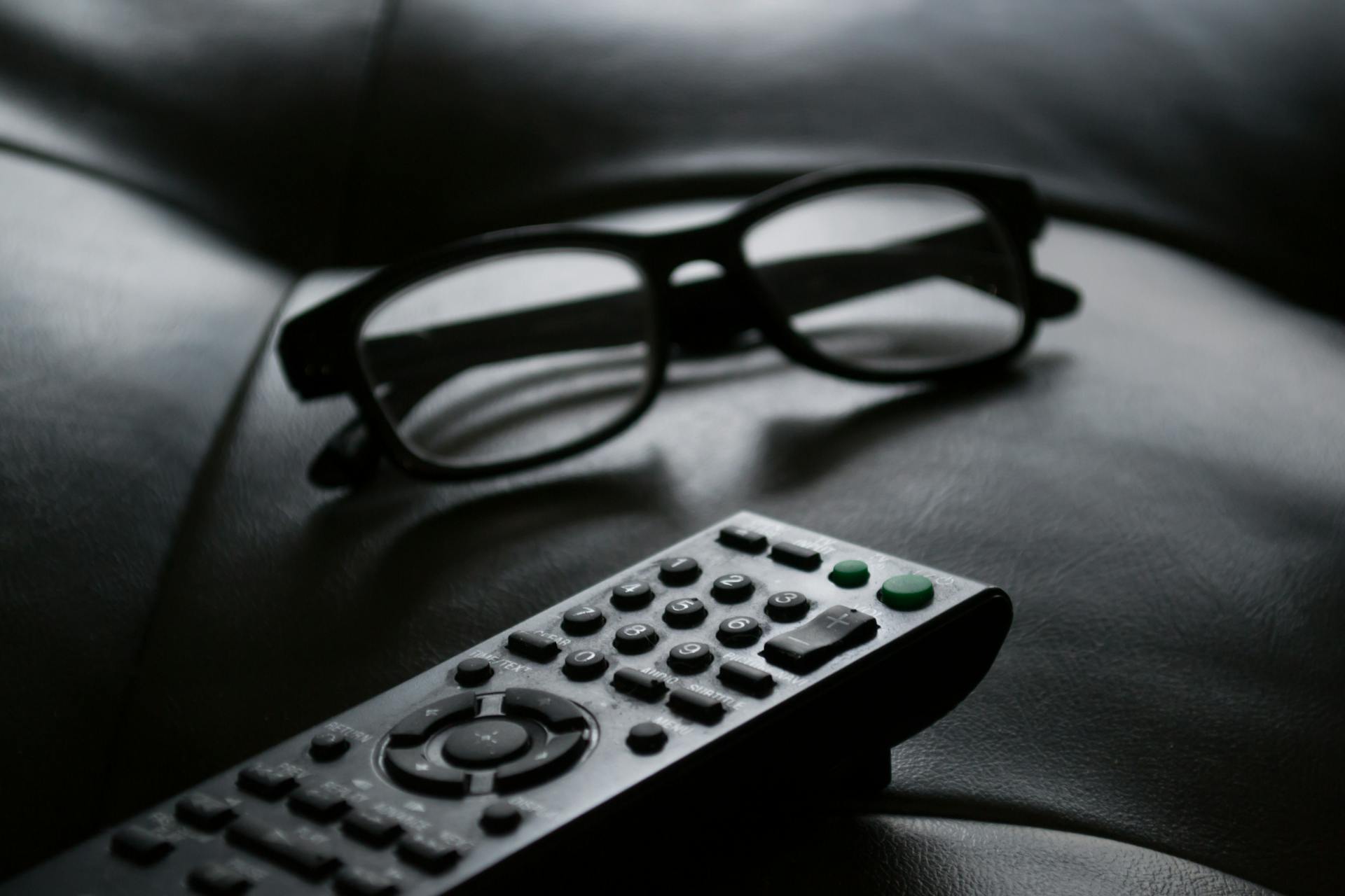 Foto en escala de grises de un control remoto cerca de unas gafas | Fuente: Pexels
