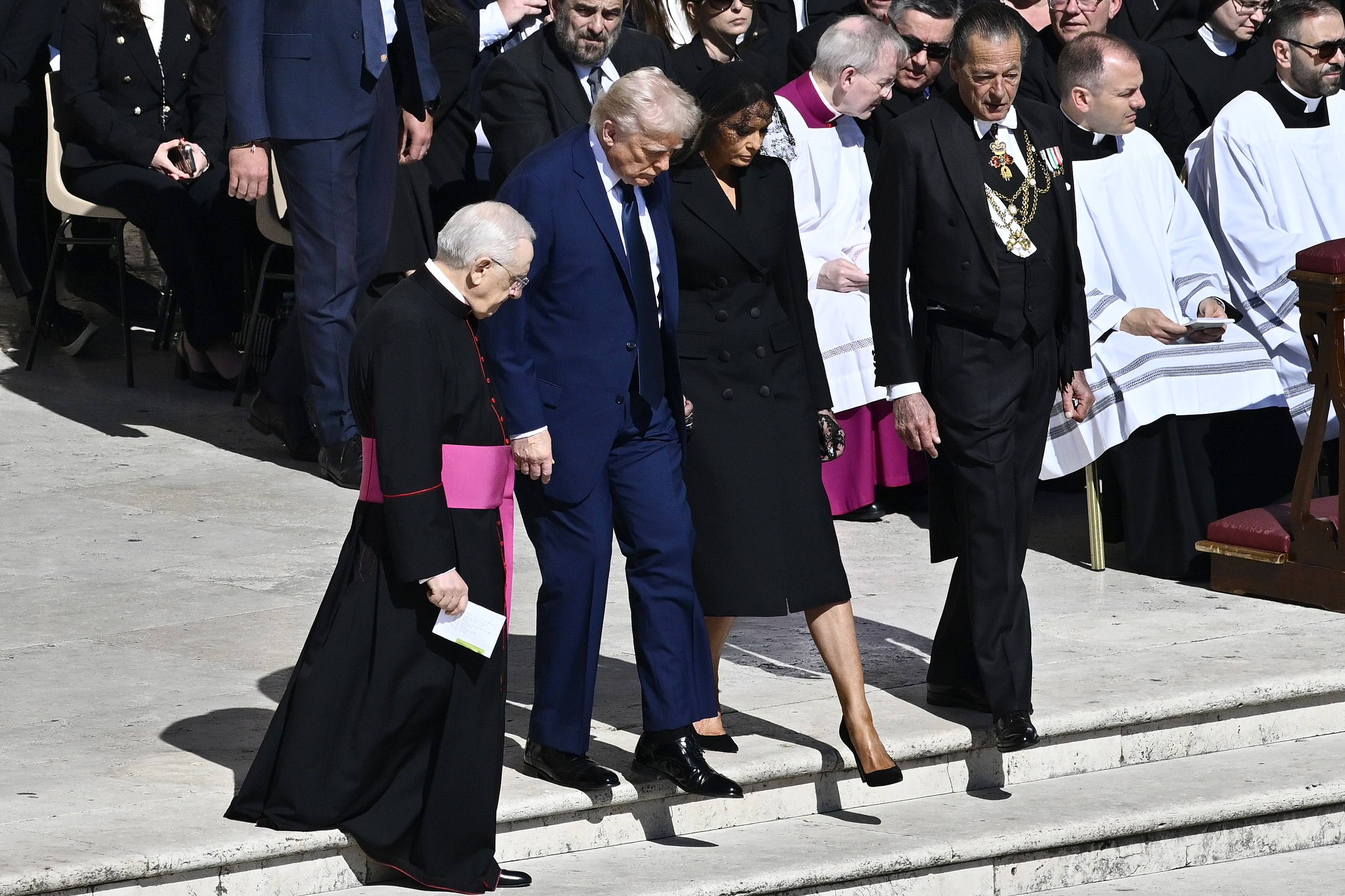 Donald y Melania Trump entran en la Plaza de San Pedro, acompañados por un sacerdote católico. Mientras que el sacerdote va vestido con una túnica negra, al igual que Melania con su atuendo, el traje azul de Donald destaca en marcado contraste, llamando la atención en medio del mar de atuendos oscuros.