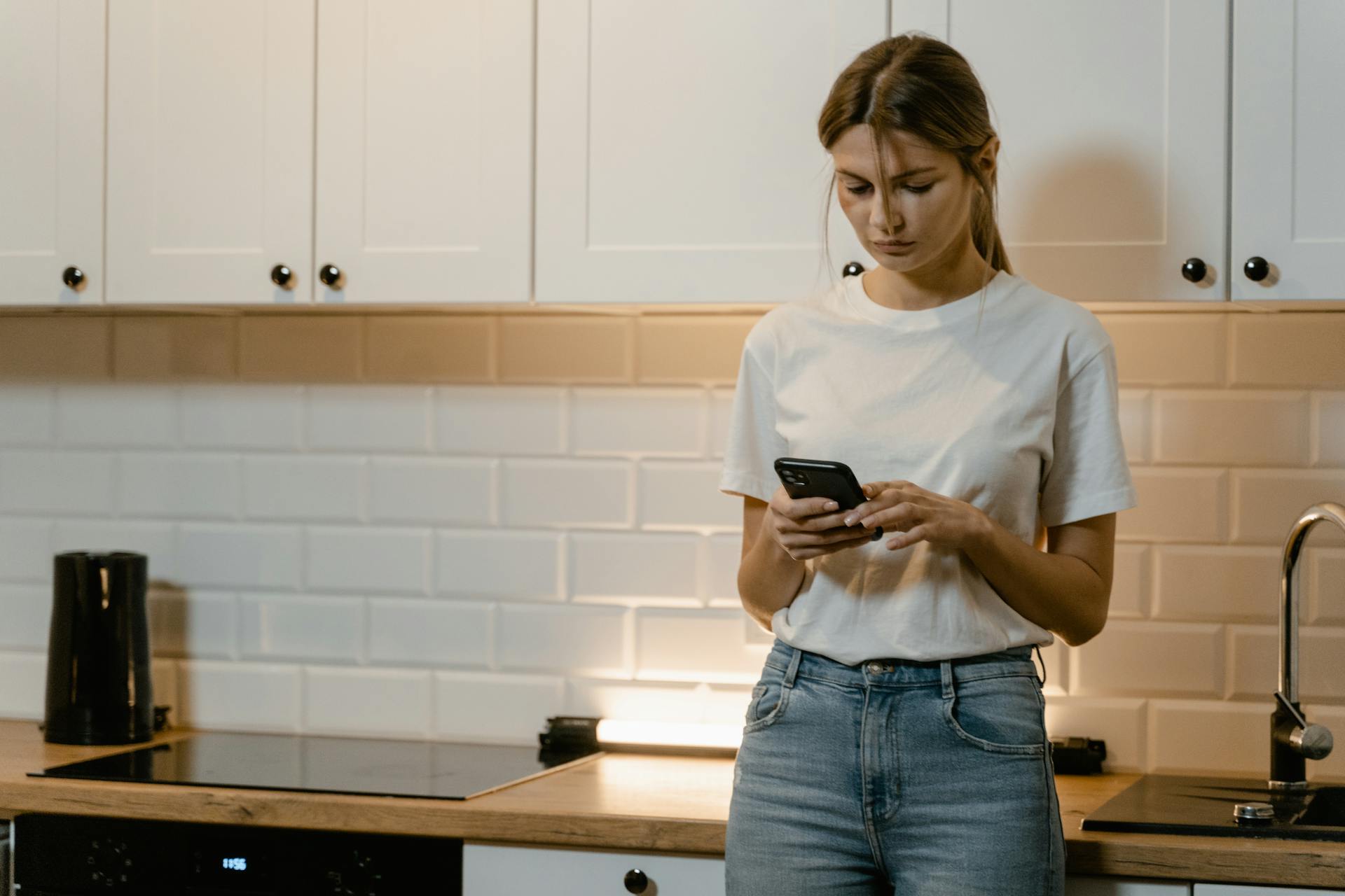 Una mujer con camiseta blanca y vaqueros utilizando un teléfono mientras está de pie en una cocina | Fuente: Pexels