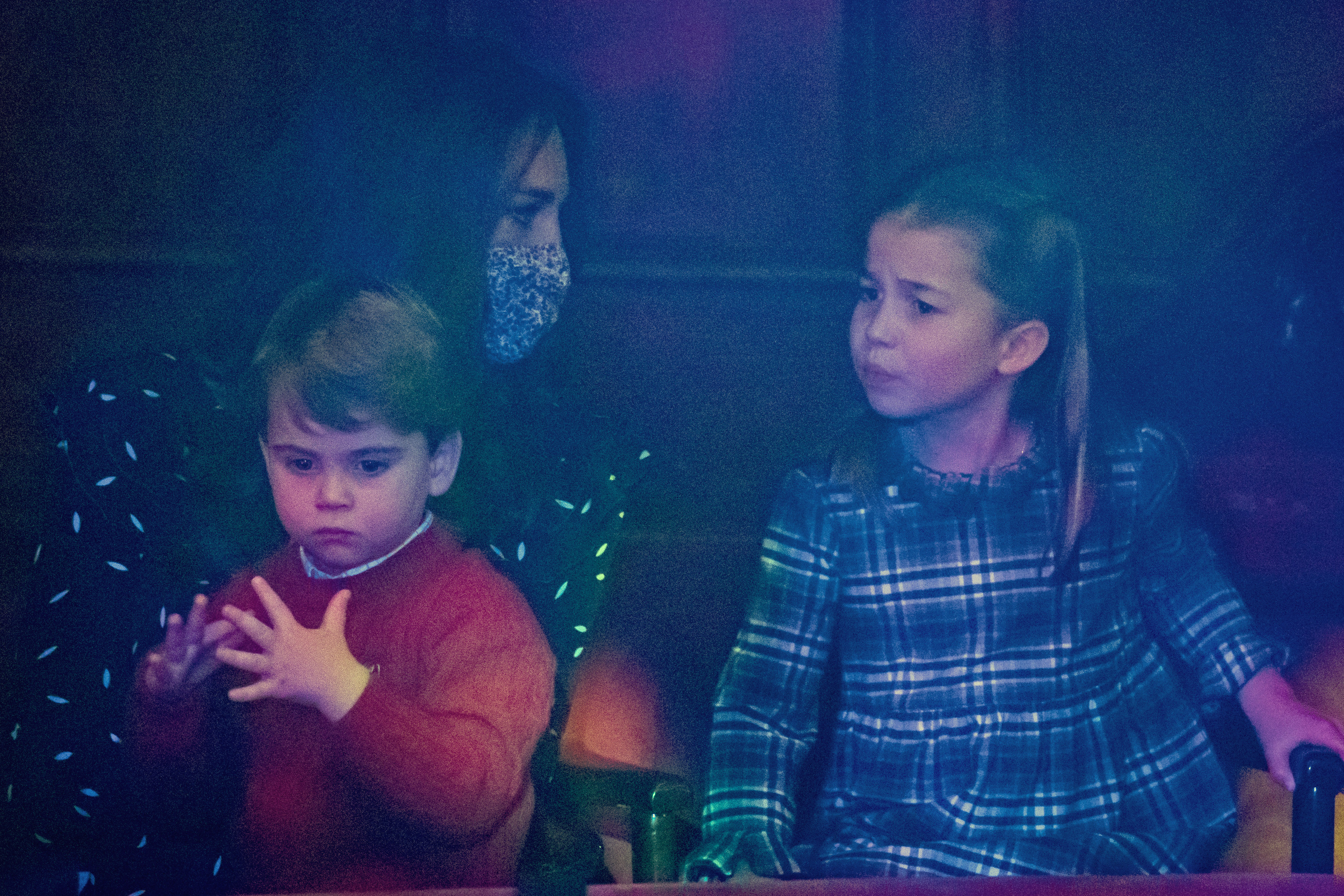 La princesa Charlotte y el príncipe Louis observan desde el público durante una representación especial de pantomima en el Teatro Palladium de Londres el 11 de diciembre de 2020, en Londres, Inglaterra, organizada para agradecer a los trabajadores esenciales y a sus familias su esfuerzo durante la pandemia. Charlotte está sentada muy atenta con un vestido de tartán azul y verde, junto a Louis, que lleva un cómodo jersey rojo de cuello redondo sobre una camisa con cuello, mientras su madre, Catherine, duquesa de Cambridge, los observa con un vestido estampado y una mascarilla protectora. | Fuente: Getty Images