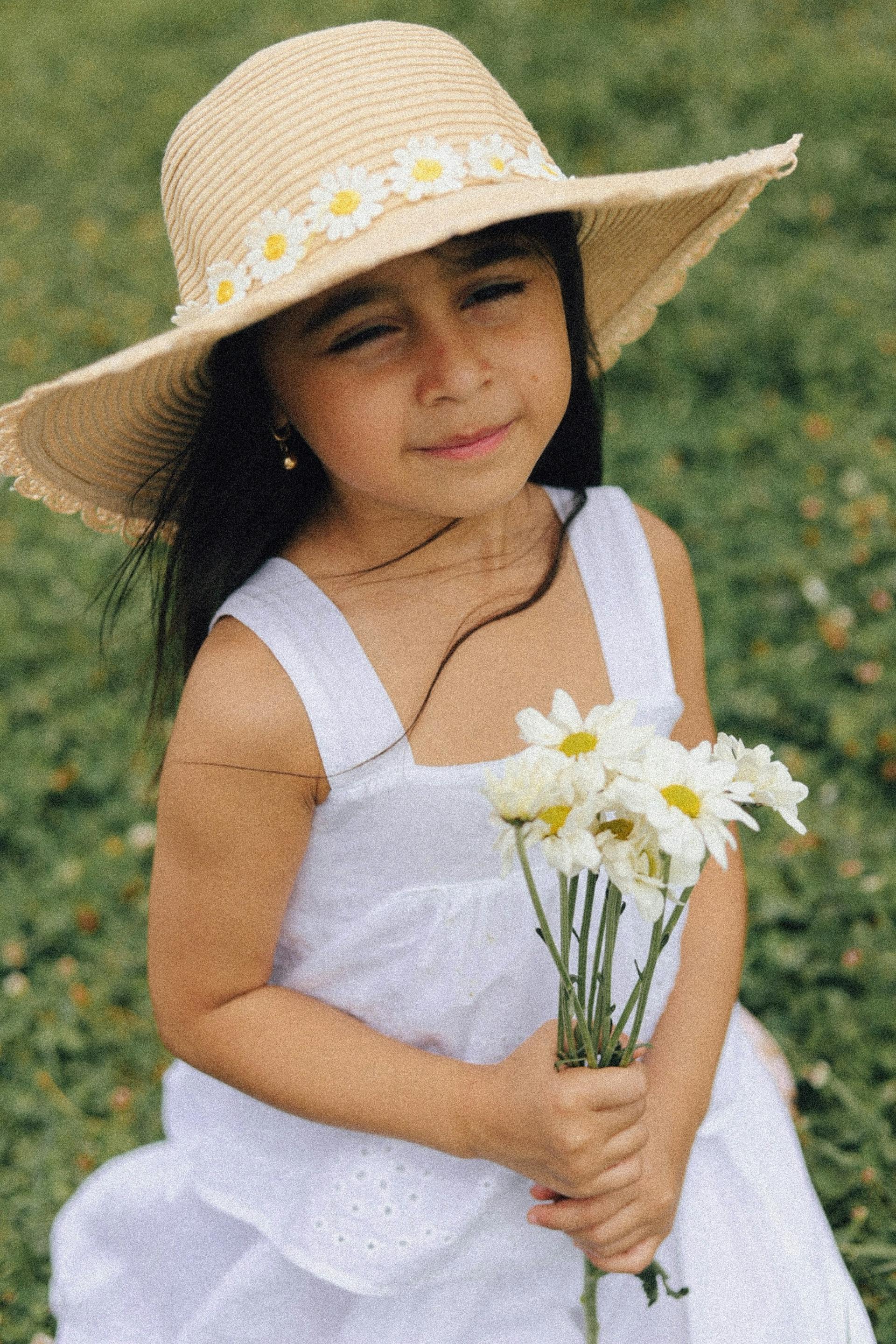 Una niña con un vestido blanco y un sombrero de paja con margaritas sujetando un ramo de flores | Fuente: Pexels