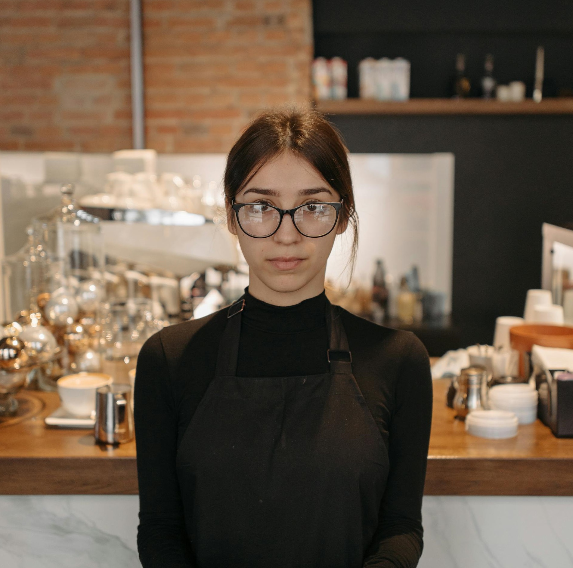 Una camarera con gafas y delantal detrás del mostrador de una cafetería | Fuente: Pexels