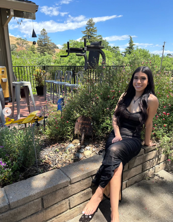 Marissa DiNapoli sentada al aire libre en una zona ajardinada, sonriendo con un vestido negro cerca de un cartel que dice "Karlene's Garden," publicado el 1 de julio de 2025 | Fuente: Instagram/moniice__