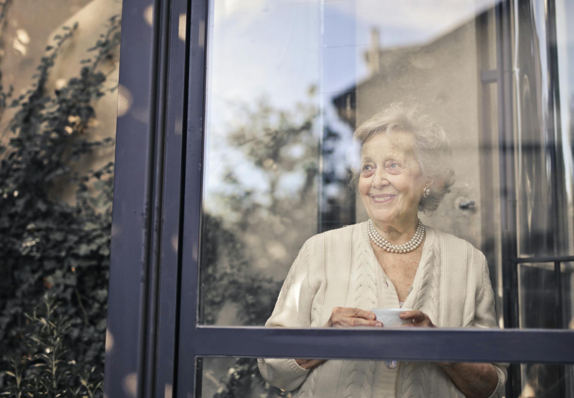 Una anciana sonriente con su taza de té en la mano y mirando por la ventana | Fuente: Pexels