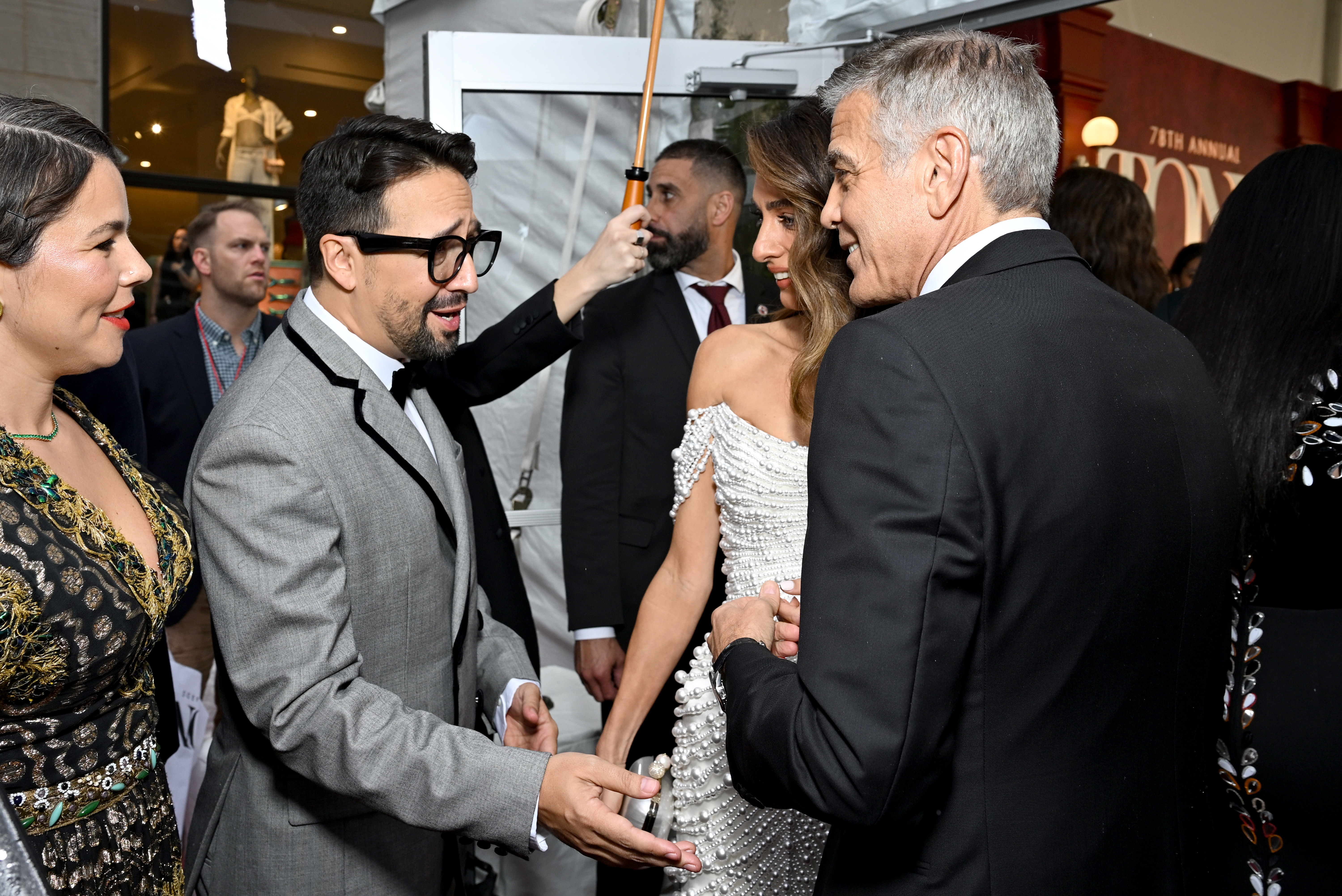En un intercambio sincero, George Clooney y Amal Clooney entablan conversación con Lin-Manuel Miranda. Amal sonríe cálidamente mientras George charla animadamente, creando un momento animado bajo el toldo rojo.