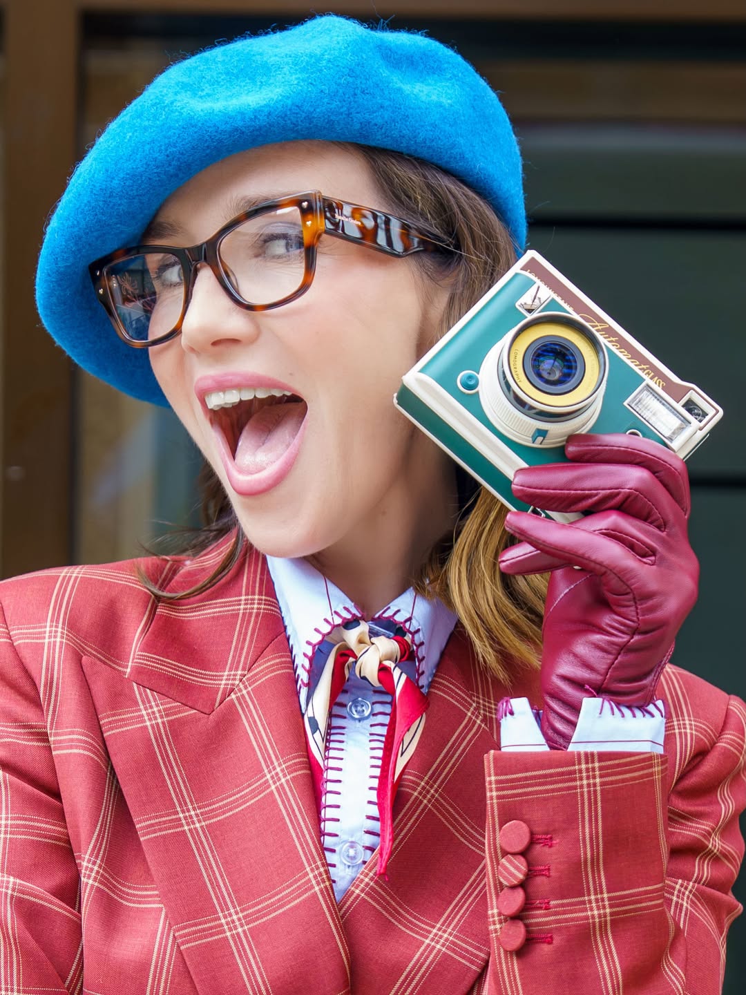 Primer plano de Natalia Oreiro con boina azul, gafas de marco carey y guantes burdeos mientras sostiene una cámara instantánea. La actriz luce su traje de cuadros rojizos y camisa celeste durante la promoción de “Nada entre los dos”. | Fuente: Instagram/nataliaoreirosoy