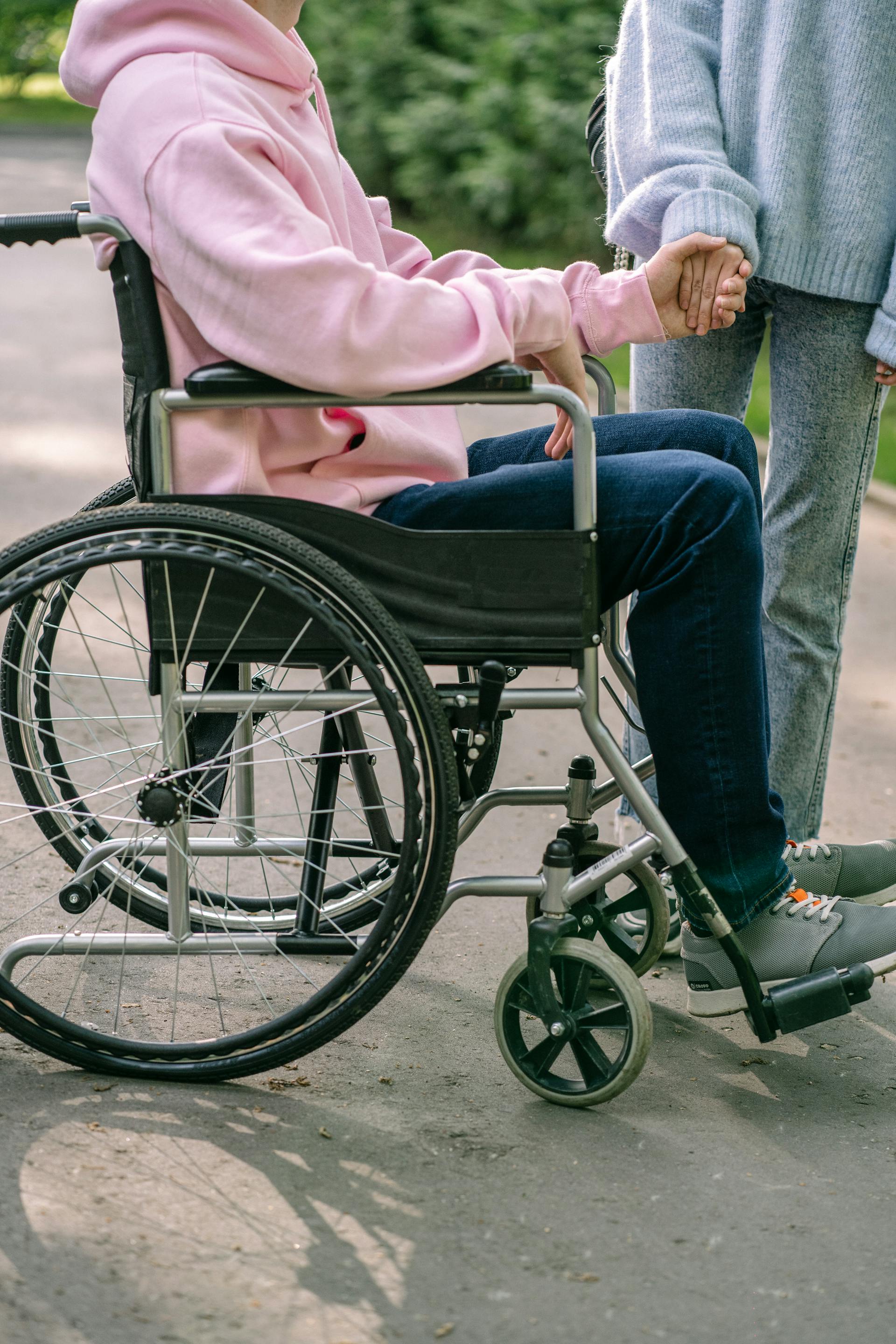 Un hombre con una sudadera rosa sentado en una silla de ruedas y cogido de la mano de una mujer | Fuente: Pexels