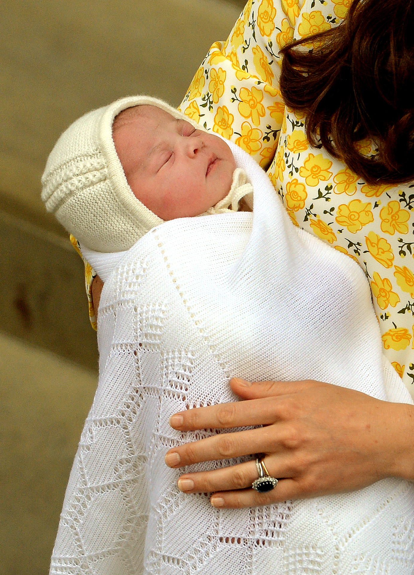 Catherine, duquesa de Cambridge, sostiene en brazos a su hija recién nacida, la princesa Charlotte, a la salida del ala Lindo del Hospital St Mary's el 2 de mayo de 2015, en Londres, Inglaterra. La pequeña, que duerme plácidamente, está envuelta en una suave manta blanca de punto con un gorrito a juego, y Catherine la sostiene con firmeza; ella lleva un vestido amarillo con estampado floral y su anillo de compromiso de zafiro. | Fuente: Getty Images