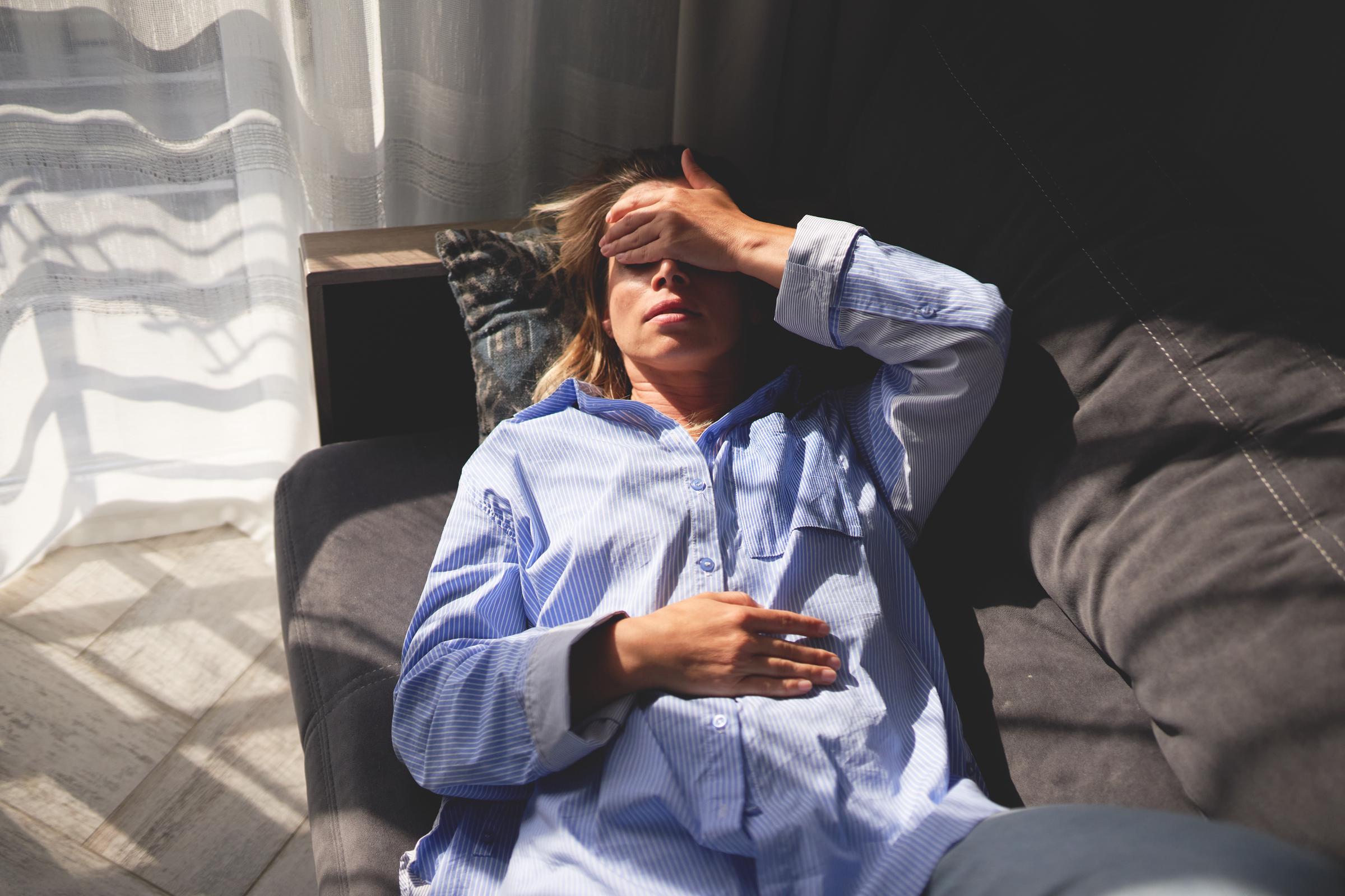 Mujer tumbada en un sofá con la mano en la frente y los ojos cerrados, lo que indica agotamiento o estrés | Fuente: Getty Images