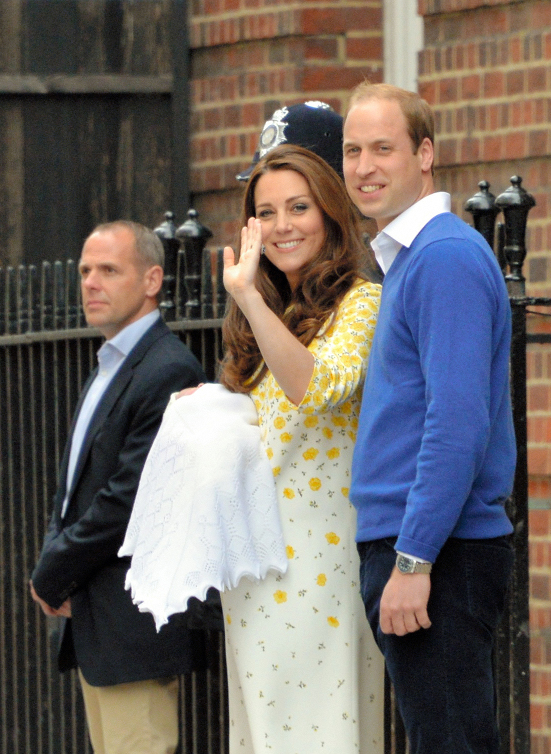 El príncipe Guillermo, duque de Cambridge, sonríe orgulloso junto a Catalina, duquesa de Cambridge, mientras ella saluda con la mano a la multitud que vitorea reunida frente a las escaleras del centro médico. Guillermo lleva un jersey de cuello en V de color azul real brillante sobre una camisa blanca de cuello abierto y pantalones azul marino oscuro, y está de pie junto a los guardias de seguridad mientras la pareja presenta al público al nuevo miembro de la familia real. | Fuente: Getty Images