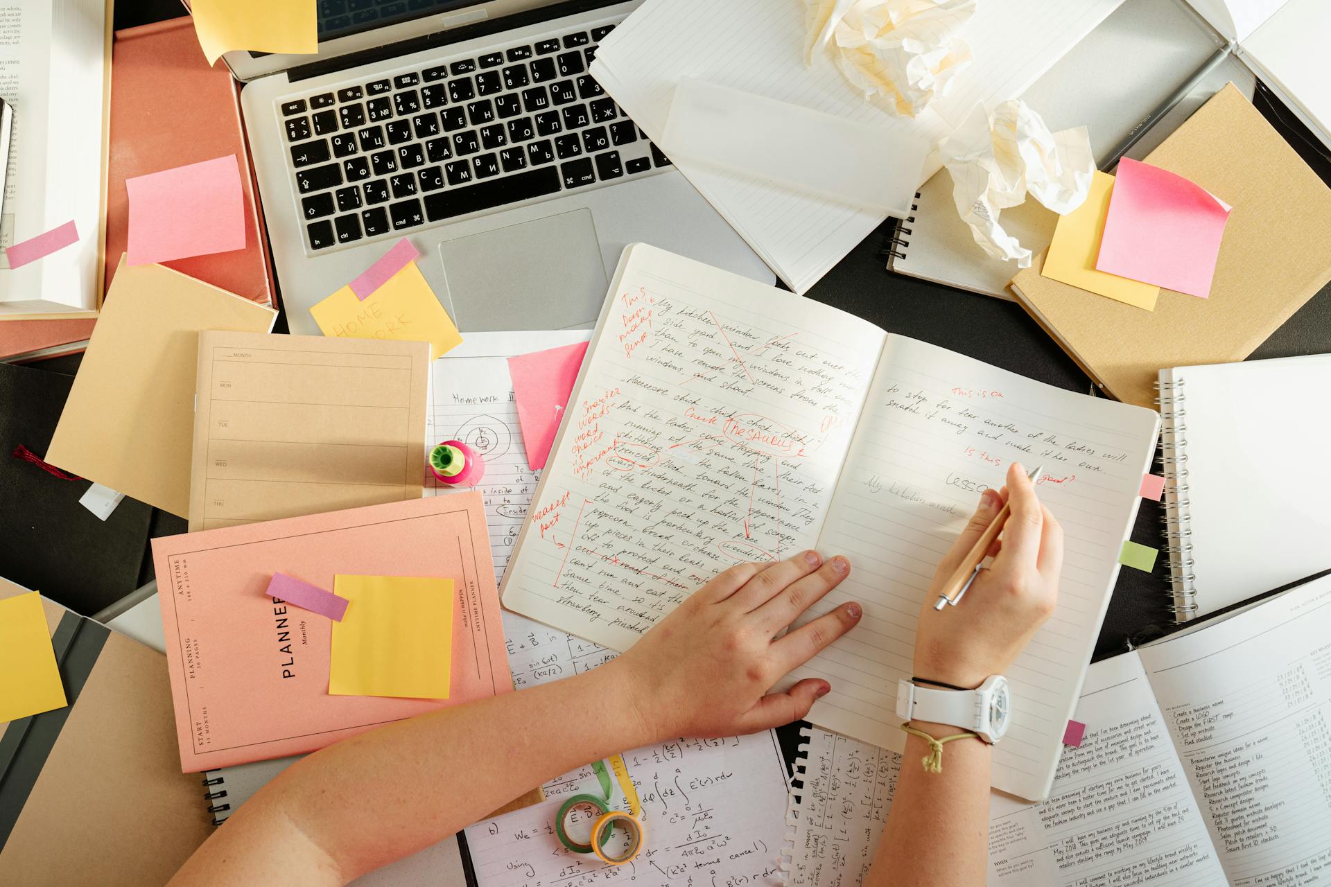 Un escritorio desordenado con libros, un portátil y una persona escribiendo en un cuaderno | Fuente: Pexels