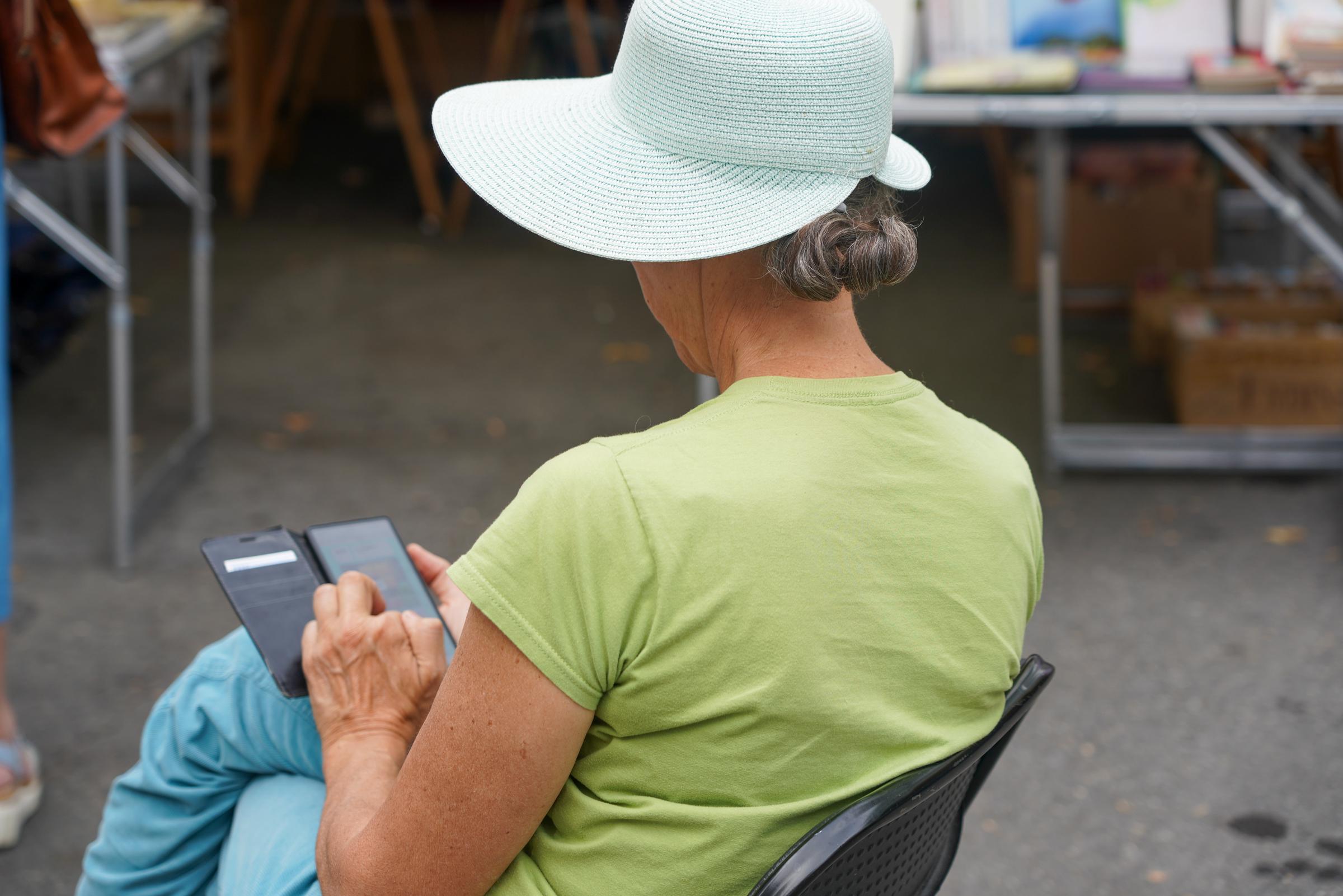 Una mujer mayor con un sombrero de ala ancha y una camisa verde sentada en una silla, usando su teléfono al aire libre. | Fuente: Getty Images