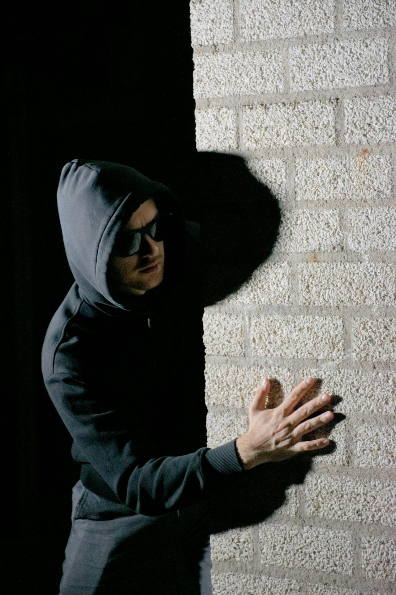 Un hombre sospechoso con capucha y gafas de sol de pie detrás de una pared | Fuente: Pexels