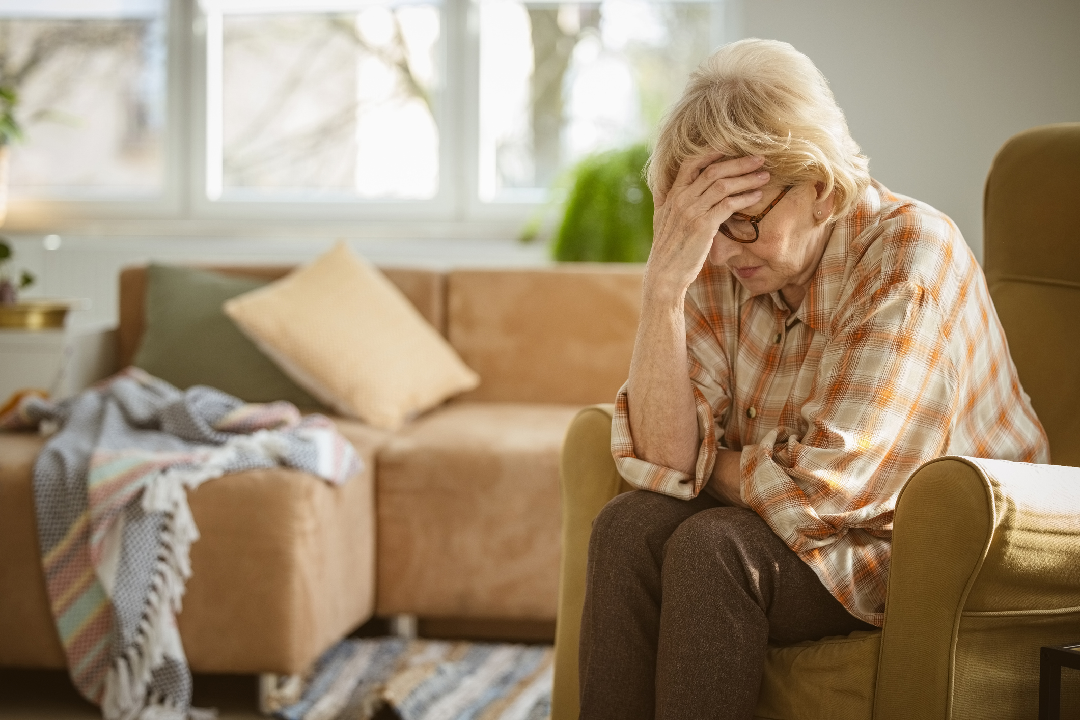 Una señora mayor preocupada y triste, con camisa a cuadros y gafas, sentada en un sillón de la sala, se toca la frente. | Fuente: Getty Images