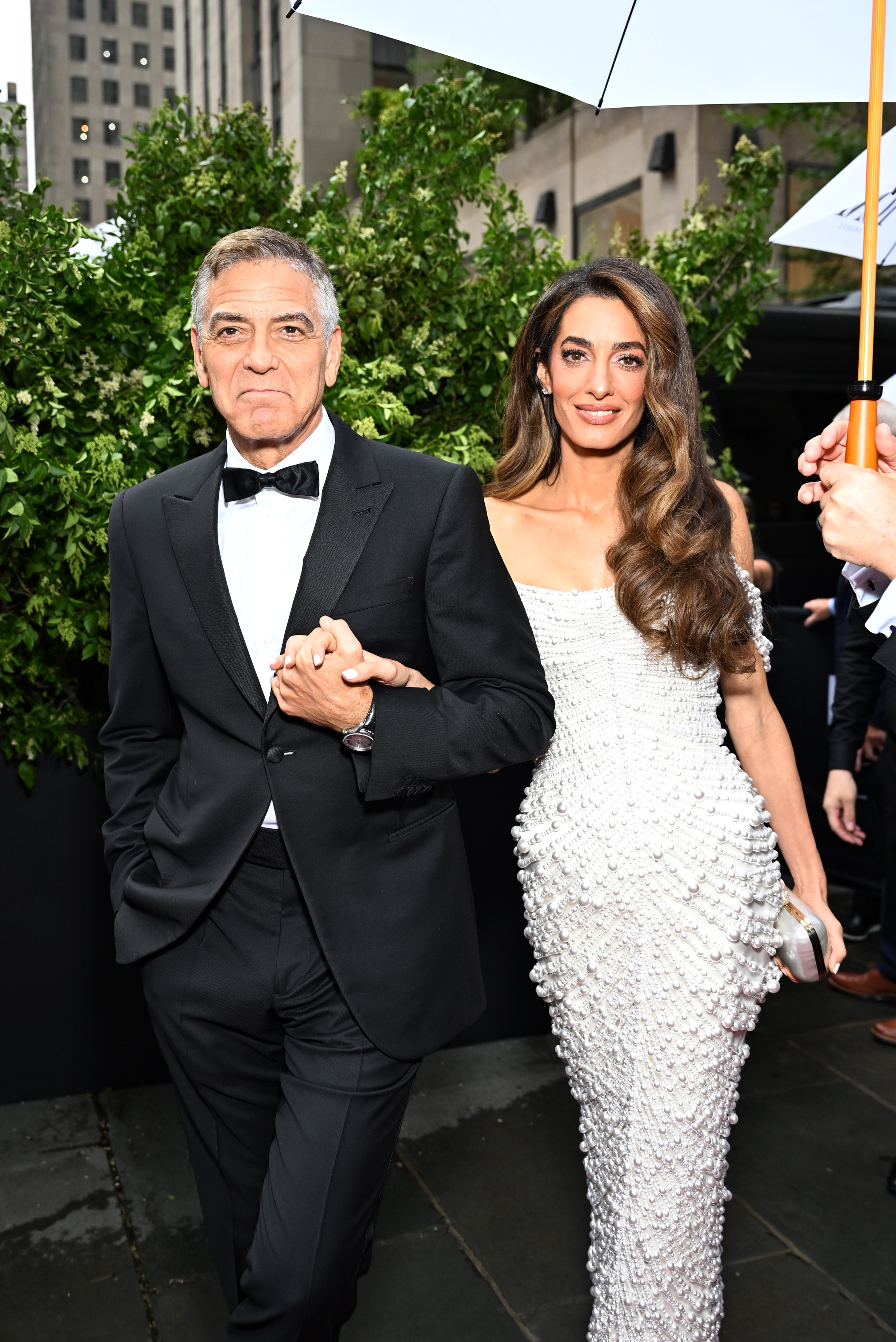 Caminando tomados del brazo, George y Amal Clooney se acercan a la entrada de la 78.ª edición de los Premios Tony, enmarcados por plantas y rodeados de personal en una noche de llovizna en Manhattan.