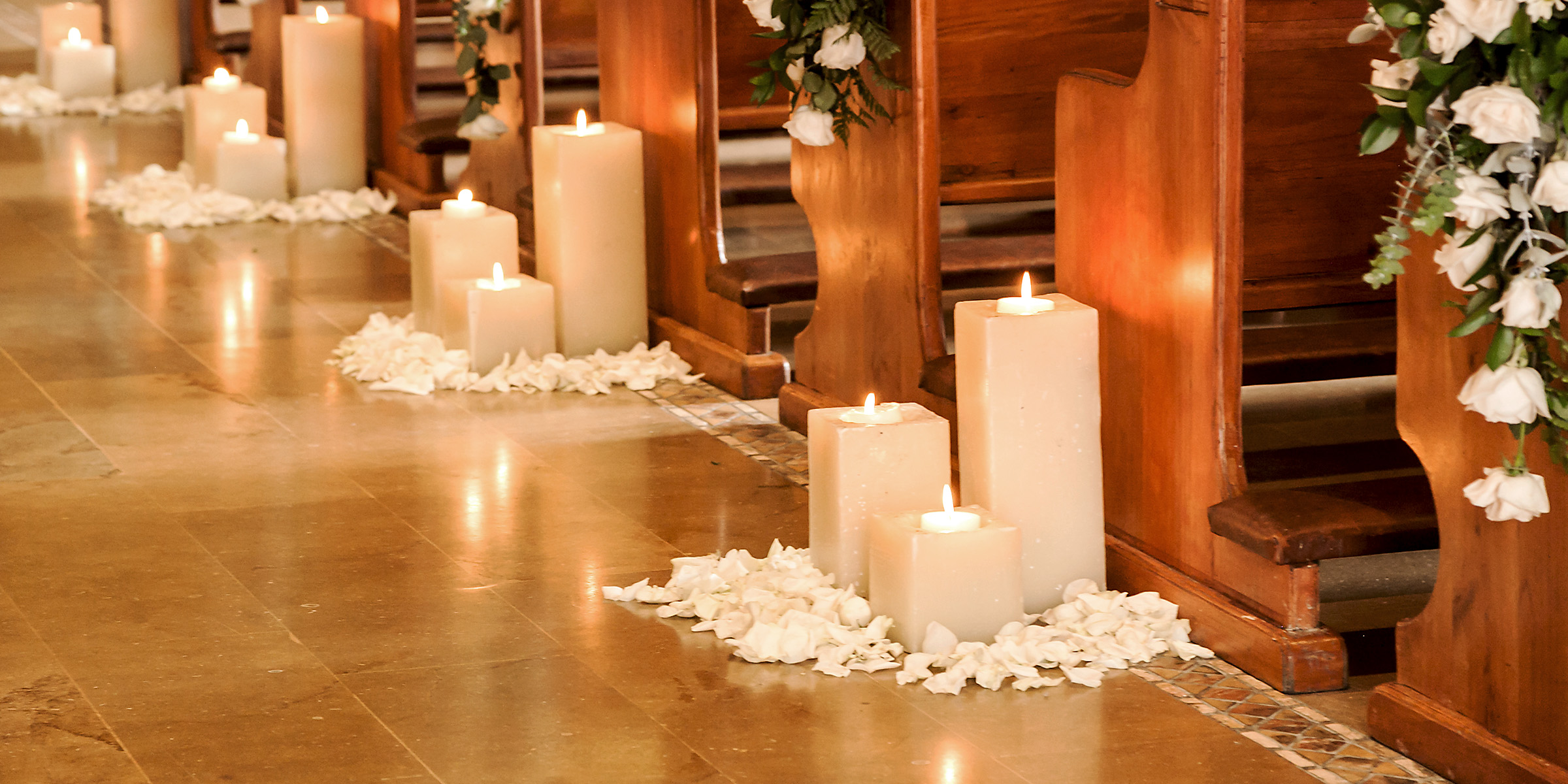 La suave luz de las velas delinea el pasillo, salpicado de pétalos de rosa blanca y enmarcado por bancos de madera | Fuente: Shutterstock
