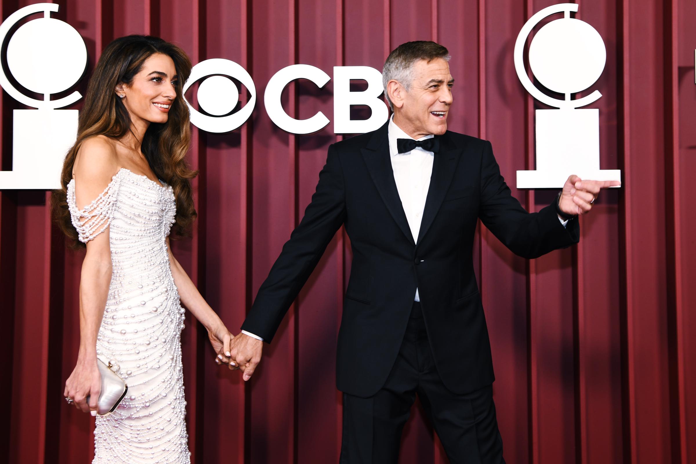 Amal Clooney resplandece mientras George Clooney charla expresivamente con sus dedos entrelazados en la alfombra roja. El vestido adornado con perlas de ella deslumbra bajo las luces, mientras que él mantiene el ambiente animado con un gesto juguetón.