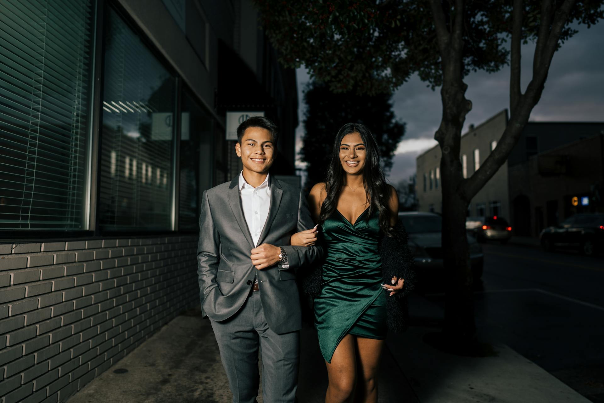 Una feliz pareja de jóvenes vestidos y caminando tomados del brazo por una calle de la ciudad por la noche | Fuente: Pexels