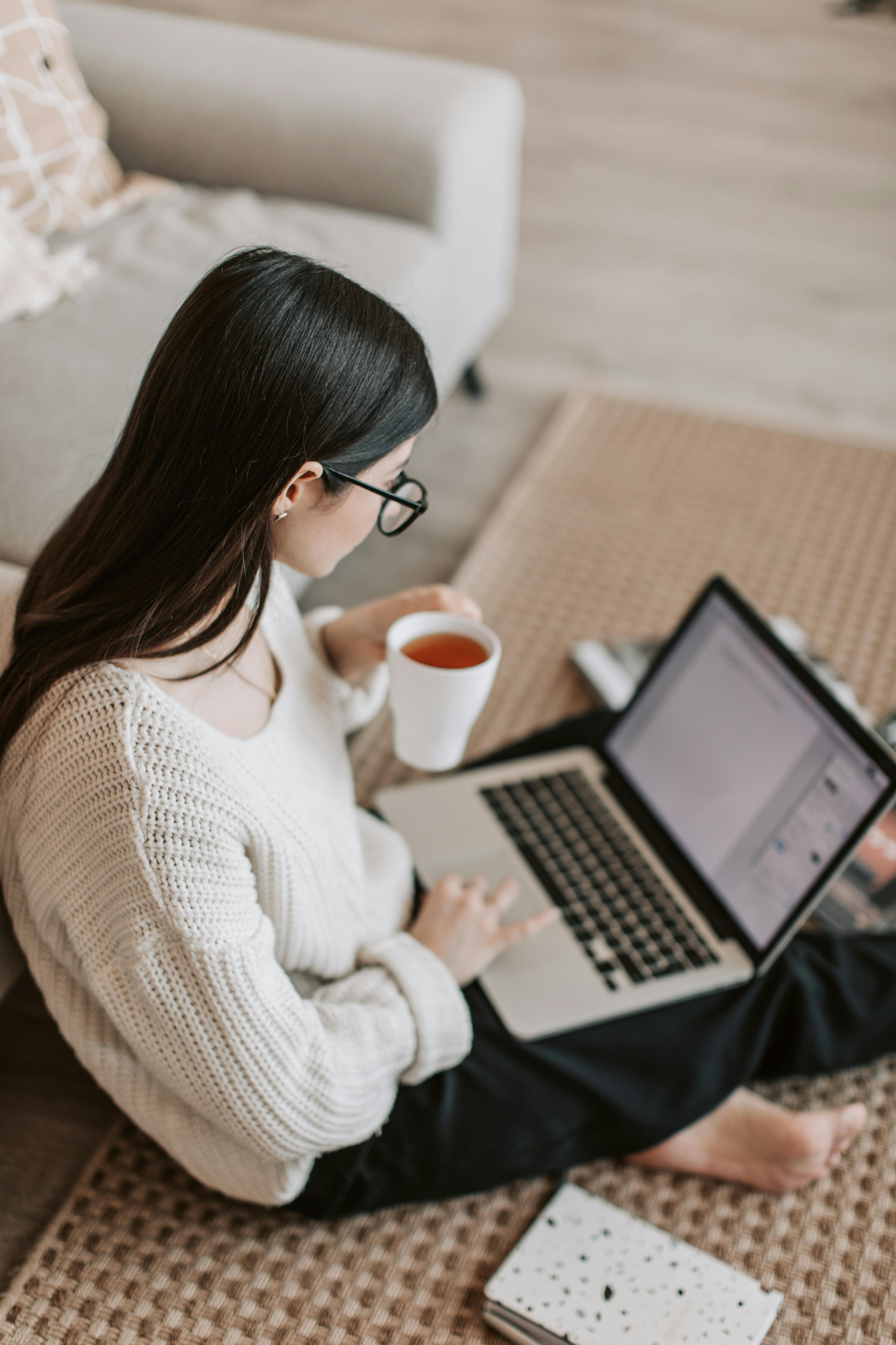 Una mujer sentada en el suelo y utilizando un ordenador portátil mientras sostiene una taza de bebida | Fuente: Pexels
