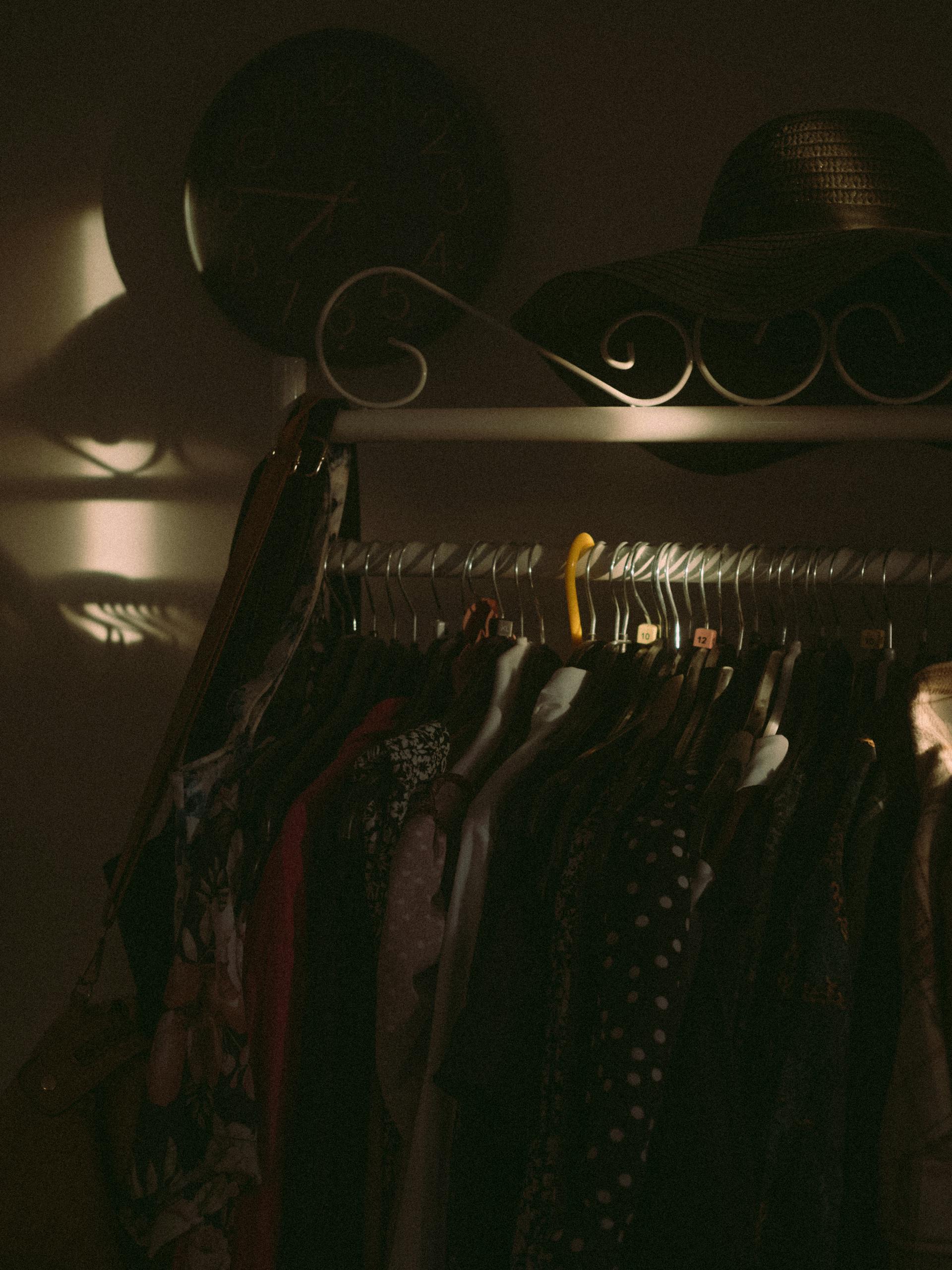 Un plano oscuro de ropa colgada en un perchero con un sombrero encima y un reloj colgado en la pared | Fuente: Pexels