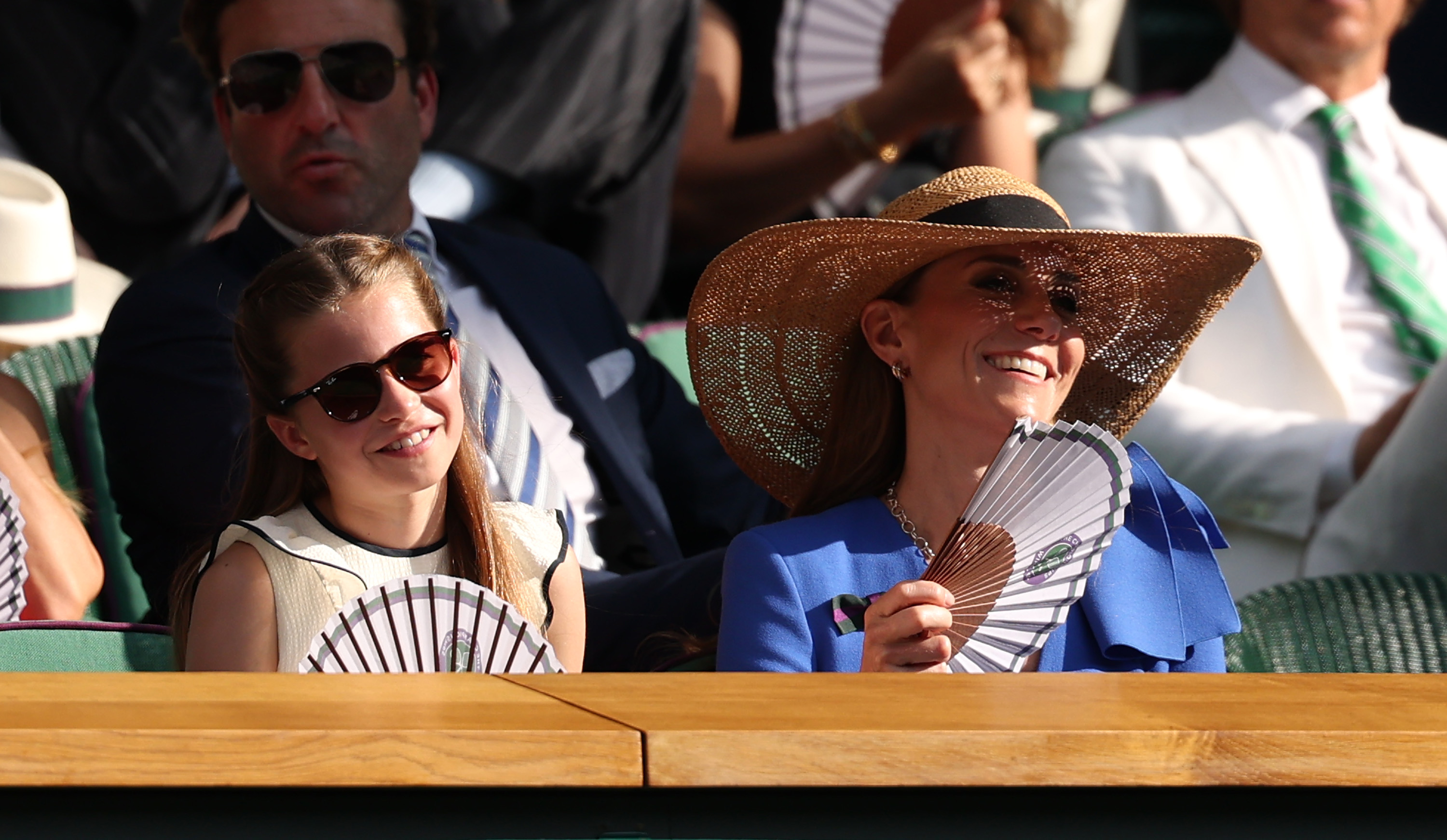 La princesa Charlotte de Gales y Catherine, princesa de Gales, sonríen mientras se refrescan con abanicos de papel en el palco real durante la decimocuarta jornada del Campeonato de Wimbledon, en el All England Lawn Tennis and Croquet Club, el 13 de julio de 2025, en Londres, Inglaterra. Charlotte ve la final individual masculina con unas gafas de sol de carey oscuro y un top sin mangas color crema con volantes, sentada junto a Catherine, que luce un look veraniego con un vestido azul brillante, unas gafas de sol llamativas y un sombrero de paja de ala ancha. | Fuente: Getty Images