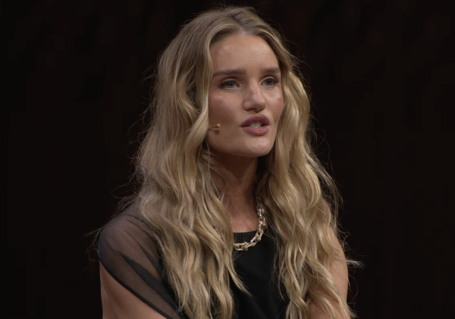 Rosie Huntington-Whiteley aparece en el escenario en plena charla, con un elegante vestido negro con detalles transparentes y un llamativo collar dorado. | Fuente: YouTube/VogueAustralia