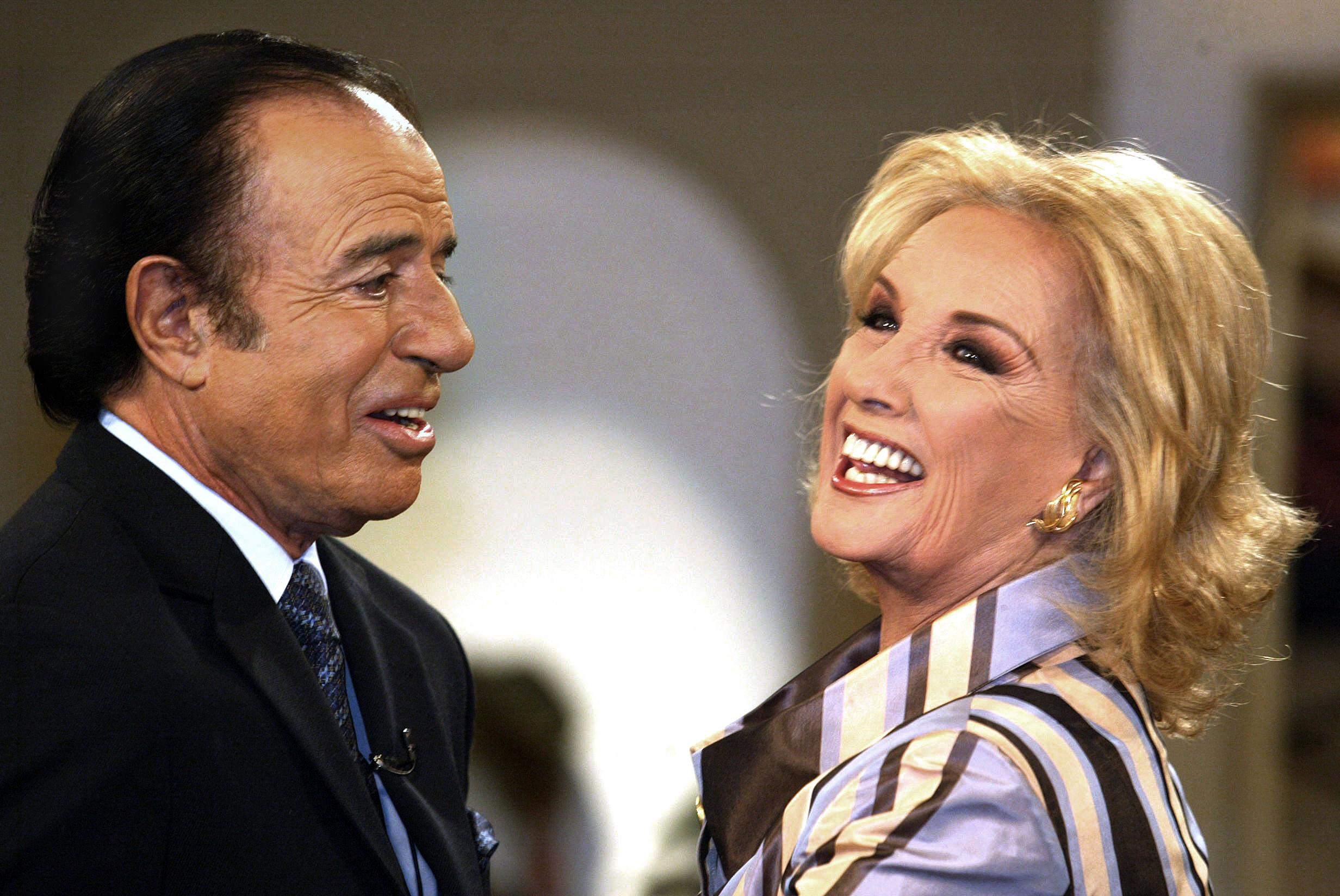Carlos Menem saluda sonriente a Mirtha Legrand al inicio del programa "Almorzando con Mirtha Legrand" en los estudios de América TV el 7 de mayo de 2003. El expresidente y candidato luce un traje oscuro con corbata estampada mientras conversa de cerca con la conductora, quien ríe con entusiasmo luciendo una blusa a rayas en tonos claros y aros dorados. | Fuente: Getty Images