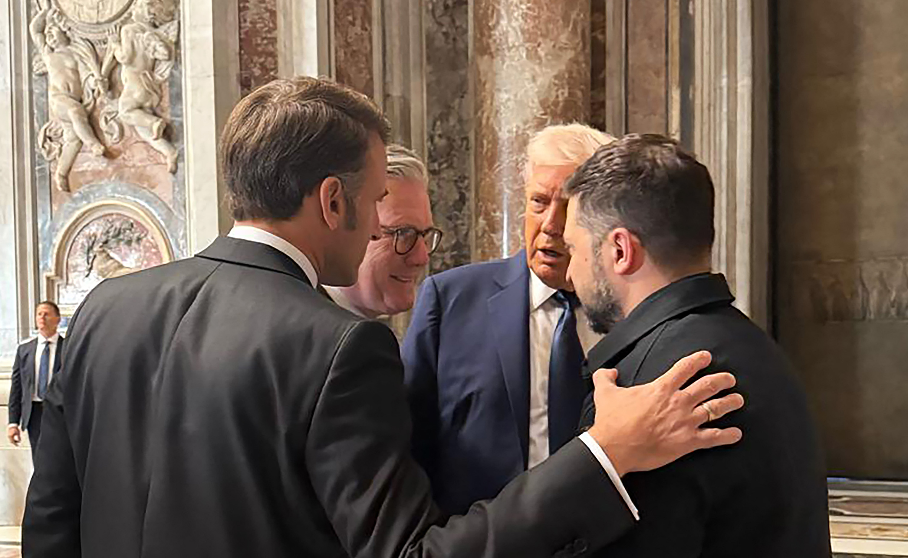En el funeral del papa Francisco, el presidente francés Emmanuel Macron pone una mano reconfortante en el hombro del presidente ucraniano Volodymyr Zelensky, ofreciéndole apoyo en medio de la guerra en curso. Junto a ellos, el primer ministro británico, Keir Starmer, y Donald, mientras conversan.