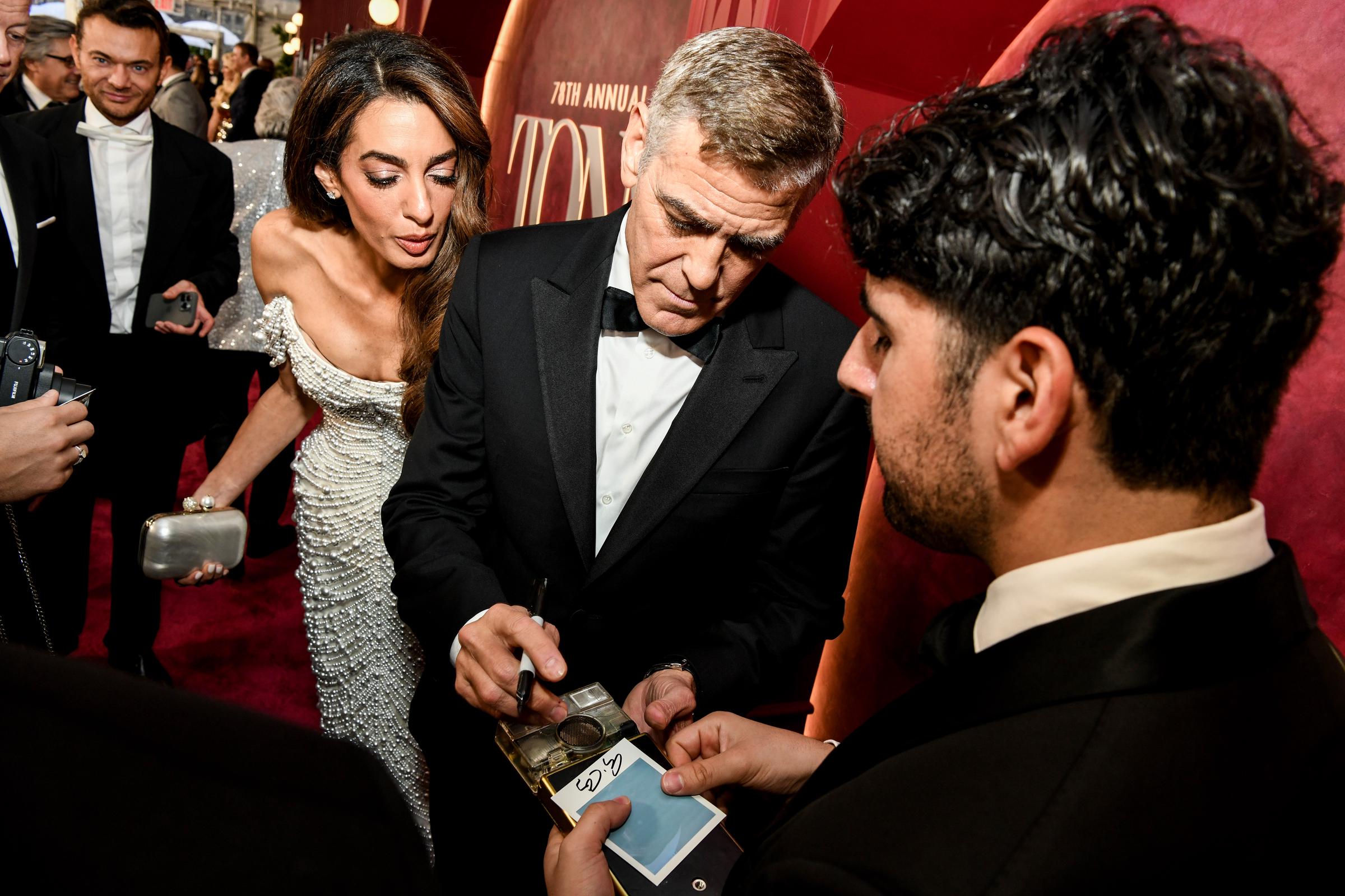 George Clooney firma autógrafos mientras Amal Clooney se inclina para observar, con expresión concentrada. La pareja hace una pausa en medio de un remolino de fans y cámaras que se disparan en la alfombra roja.