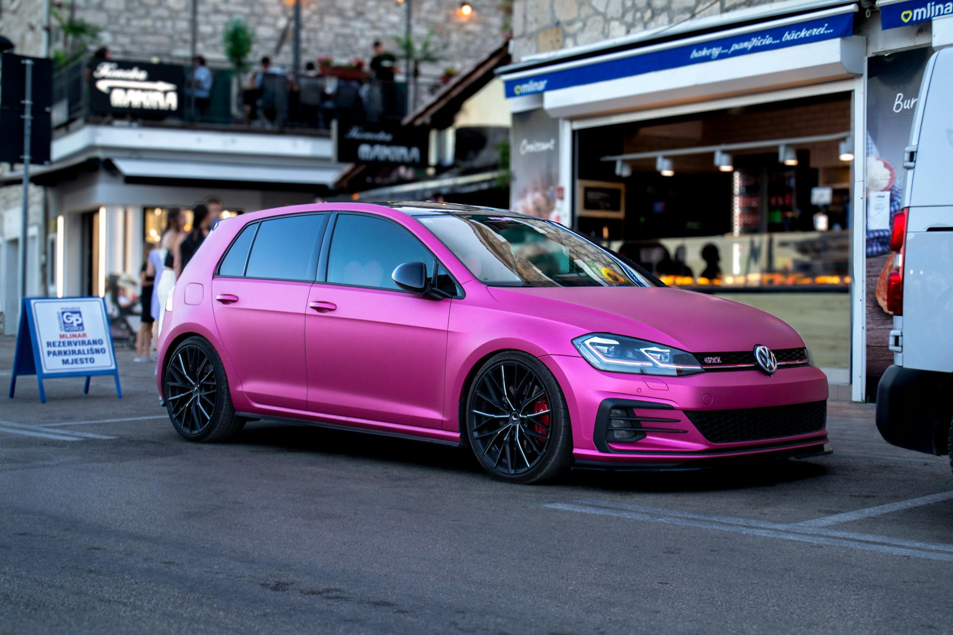 Un Automóvil con portón trasero rosa mate aparcado en una calle con edificios y gente de fondo | Fuente: Pexels