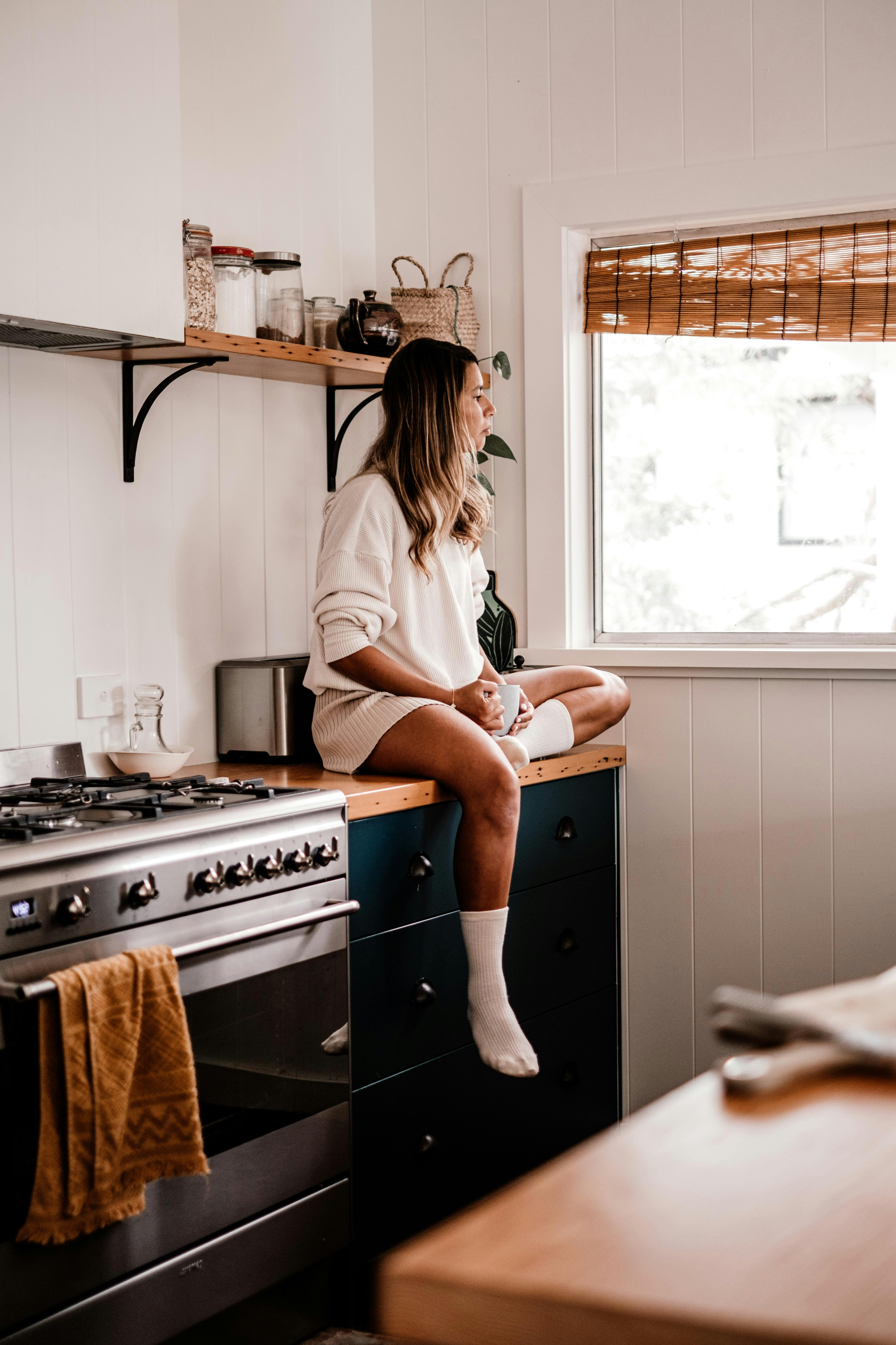 Mujer sentada en la encimera de la cocina mirando al exterior | Fuente: Pexels