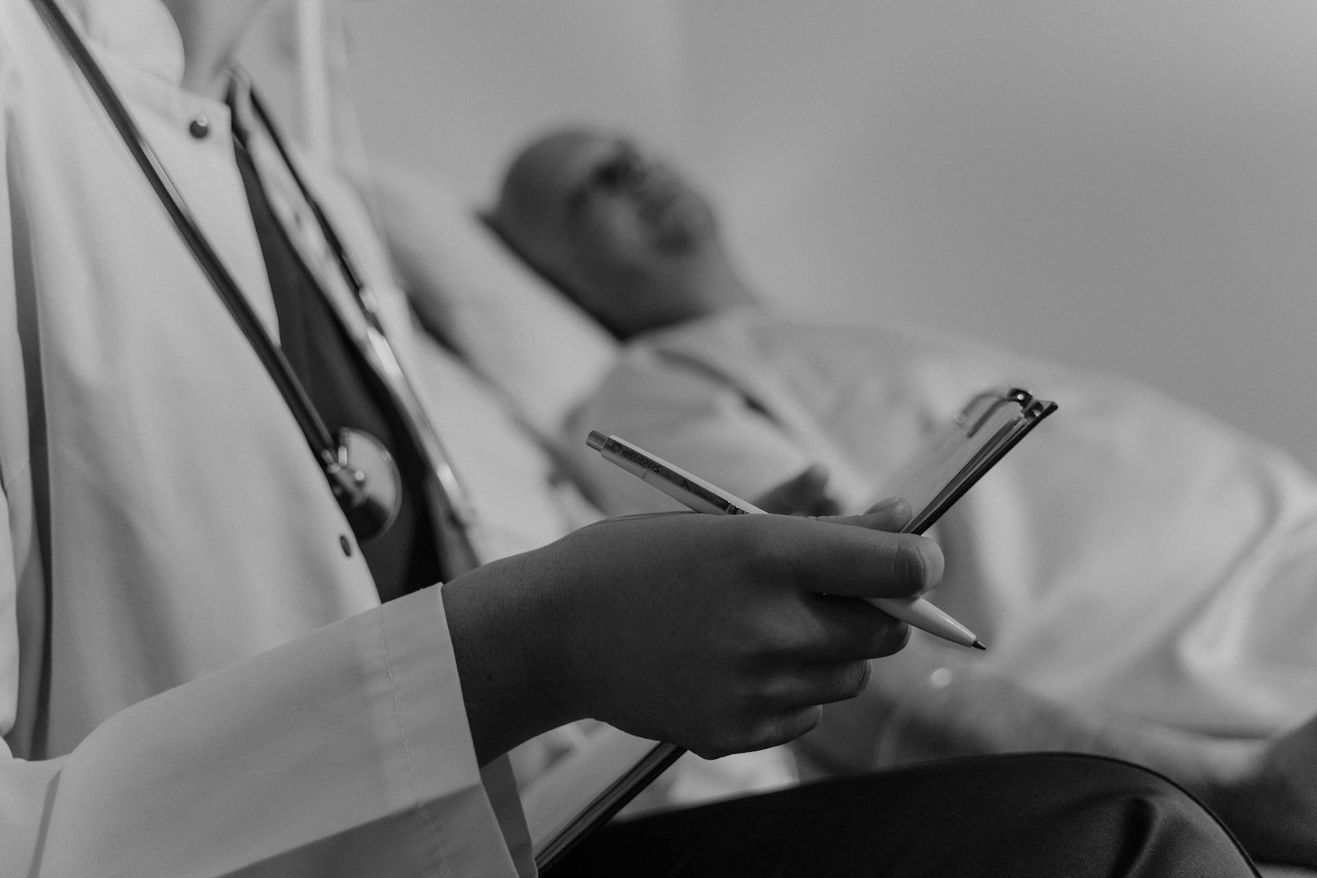 Foto en escala de grises de un médico sosteniendo un historial médico mientras está de pie junto a un paciente | Fuente: Pexels