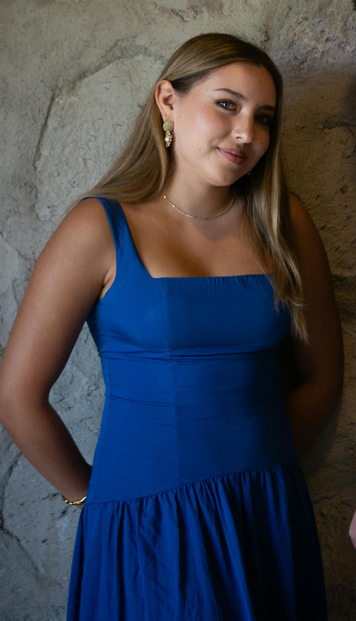 Una joven sonriente con un vestido azul, un collar y unos pendientes, de pie frente a un muro de piedra | Fuente: Pexels