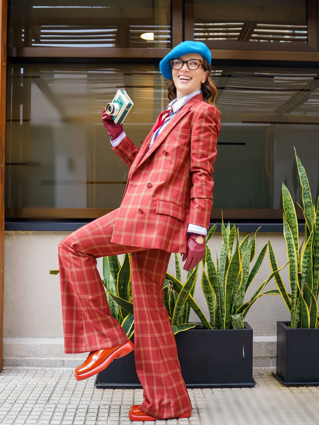 Natalia Oreiro posando al aire libre con un conjunto sastrero de cuadros en tonos rojizos. El look incluye saco y pantalón amplio, combinados con camisa celeste, pañuelo estampado, guantes burdeos, boina azul y mocasines rojos. También lleva gafas y sostiene una cámara instantánea mientras sonríe | Fuente: Instagram/nataliaoreirosoy