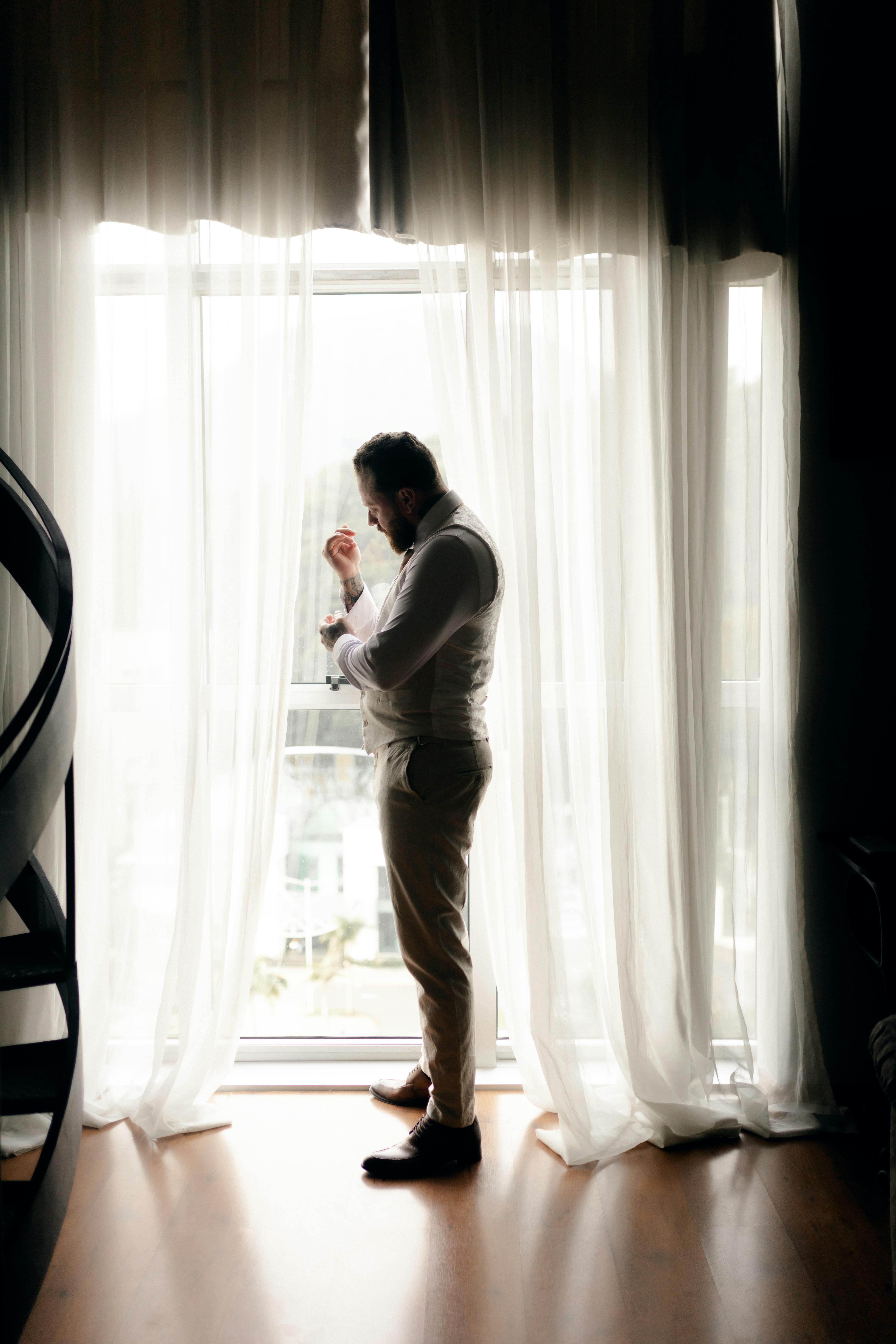 Hombre de pie junto a la ventana ajustándose el traje | Fuente: Pexels
