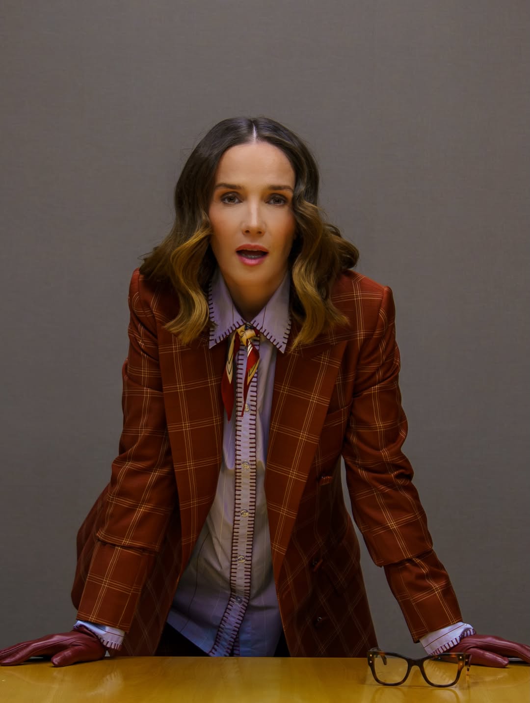 Natalia Oreiro posa apoyada sobre una mesa durante una actividad promocional de “Nada entre los dos”. La actriz lleva el traje de cuadros rojizos, camisa celeste y guantes burdeos, mientras sus gafas descansan sobre la mesa frente a ella. | Fuente: Instagram/nataliaoreirosoy